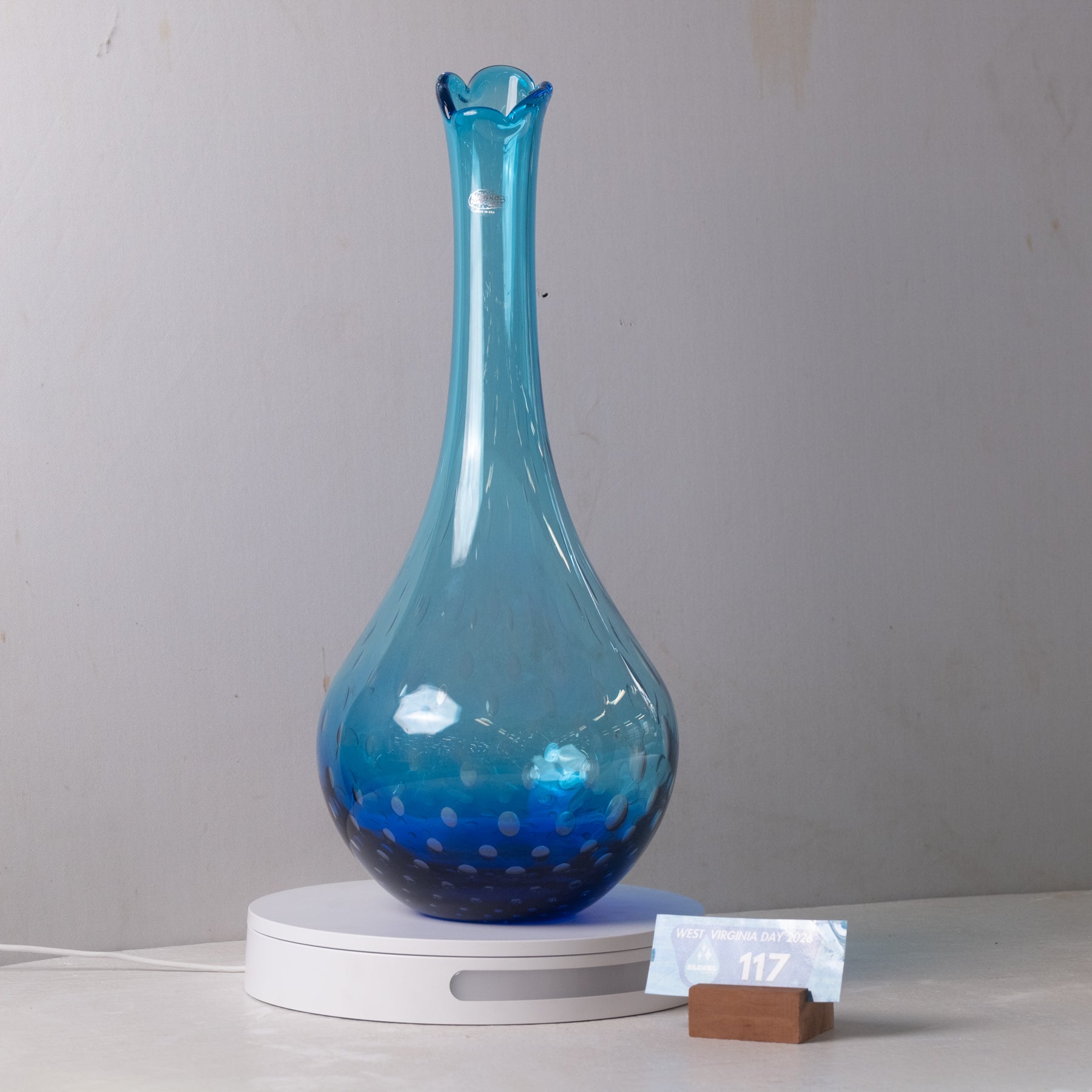 A translucent blue glass vase with a flared neck and spotted pattern near the base, displayed on a white circular light box with a small price card in front, against a plain light gray wall.