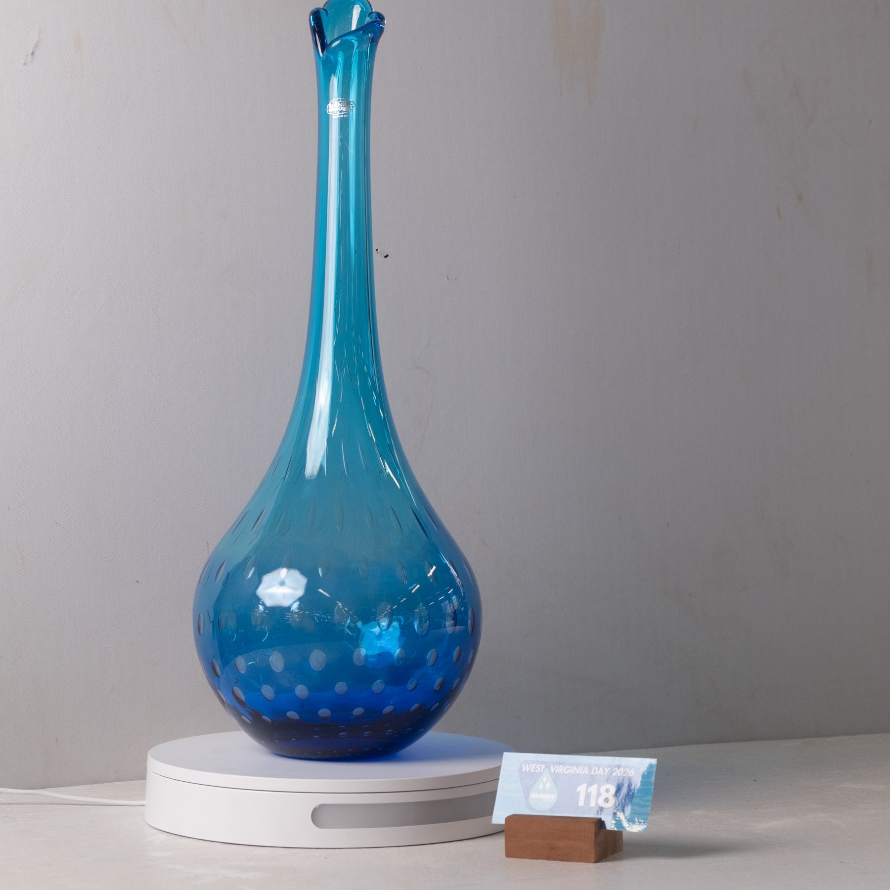 A translucent blue glass vase with a bulbous base and a long, narrow neck, displayed on a white circular light box with a small price tag in front, against a plain gray wall.