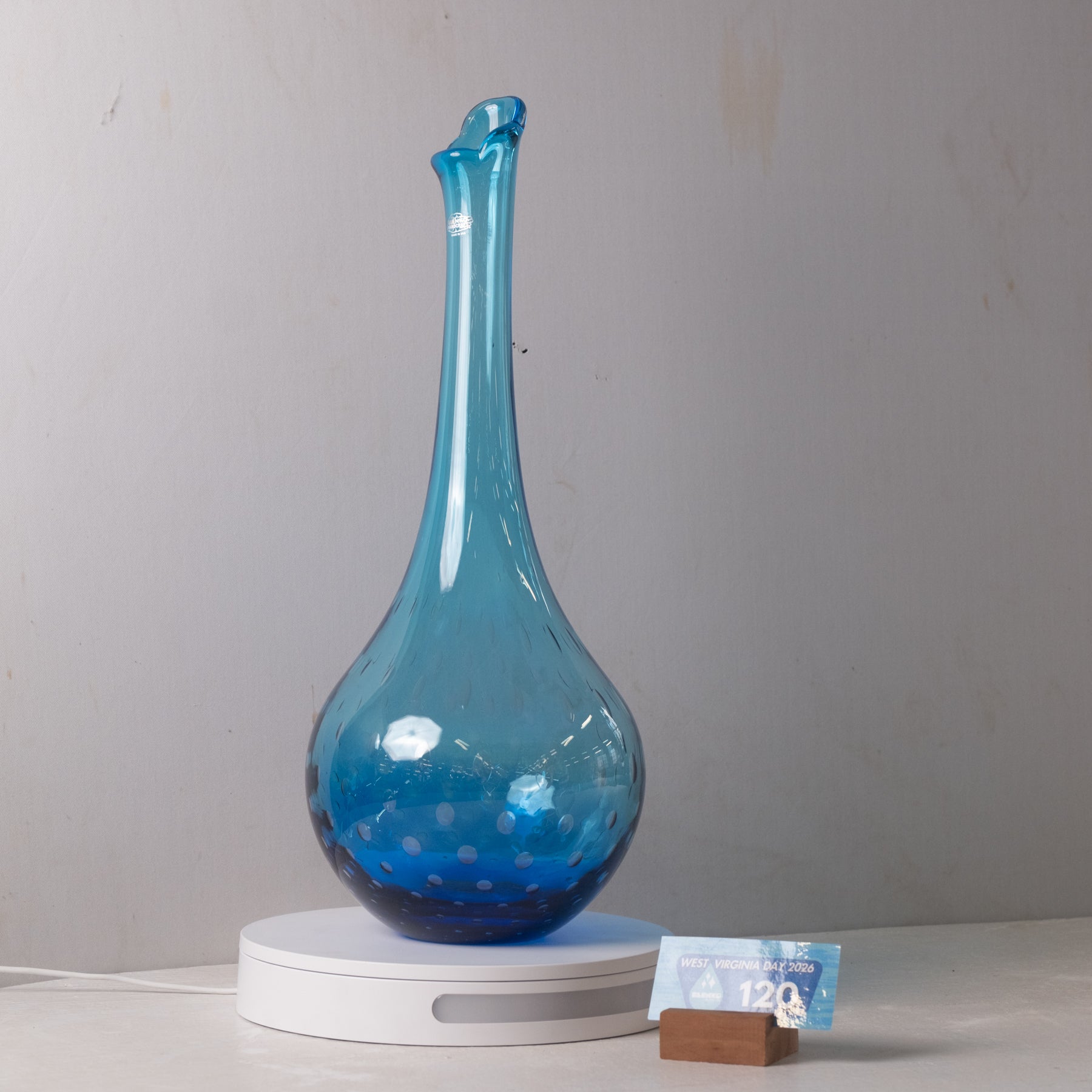 A translucent blue glass decanter with a long neck and rounded base, displayed on a white circular base against a plain light gray wall. A small Canadian 120-dollar bill is propped up next to the base as a size reference.