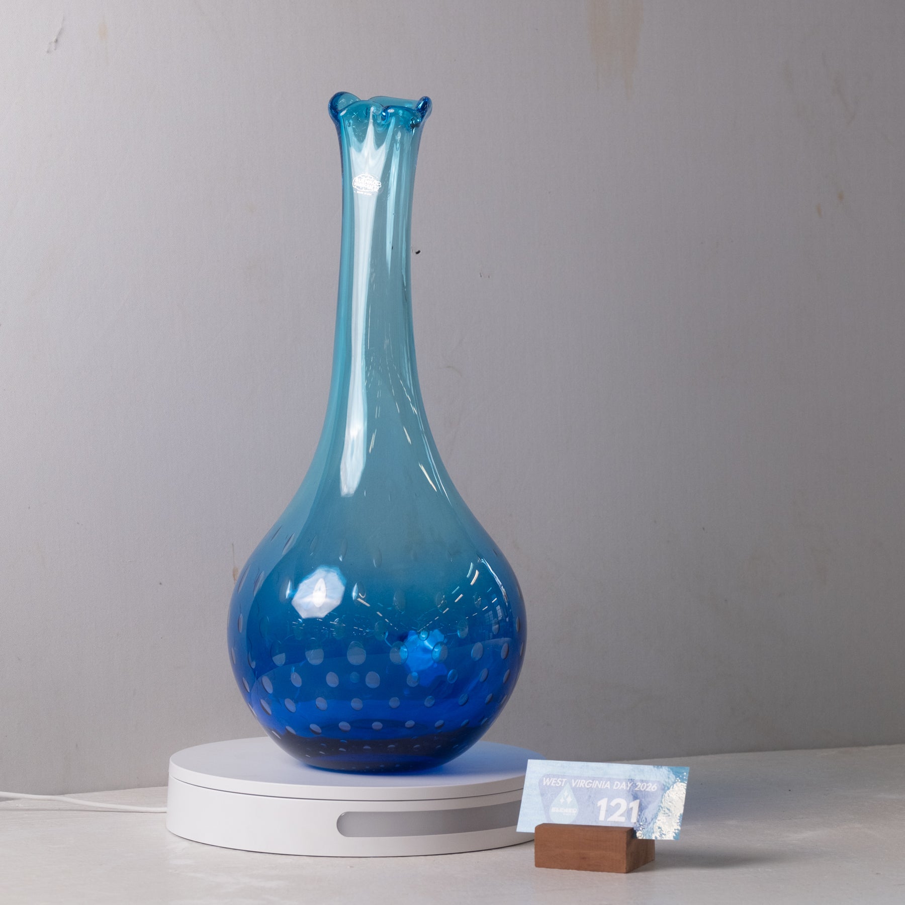 A blue glass vase with a flared neck and dotted pattern on the lower half, displayed on a white circular stand against a plain light gray wall.
