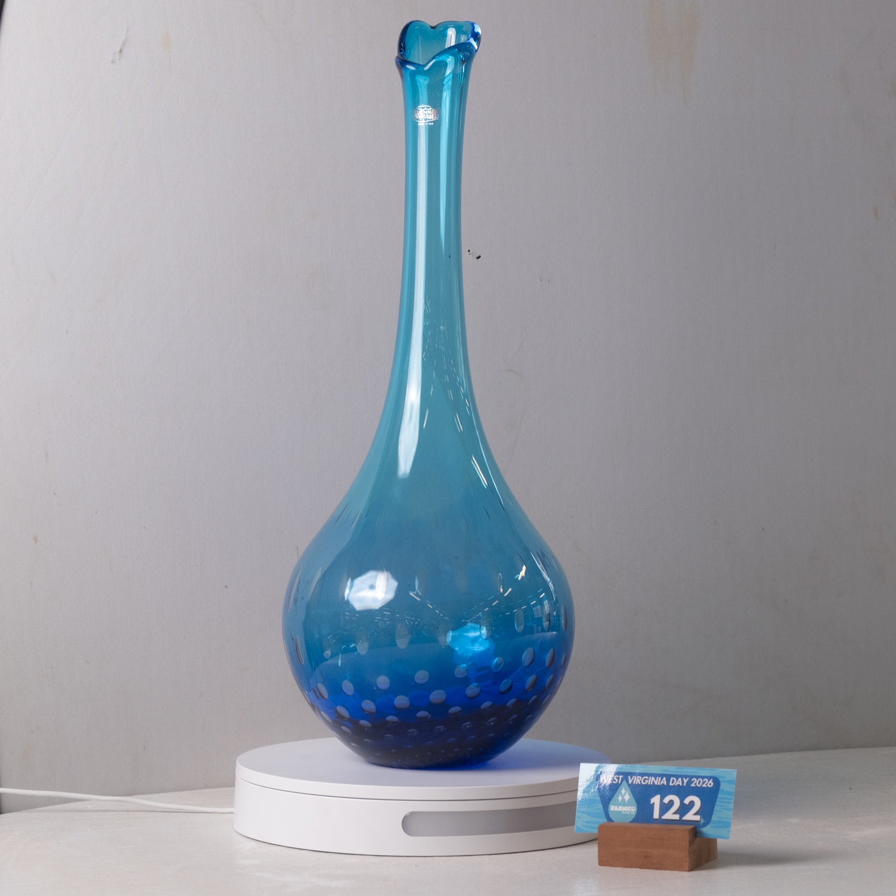 A translucent blue glass vase with a flared neck and dotted pattern near the base, displayed on a white circular light box with a small price card in front, against a plain light gray wall.