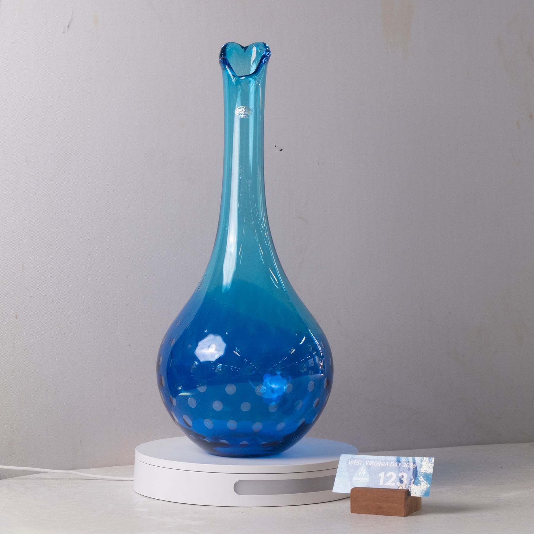 A translucent blue glass vase with a flared neck and dotted pattern near the base, displayed on a white circular stand against a light gray wall.