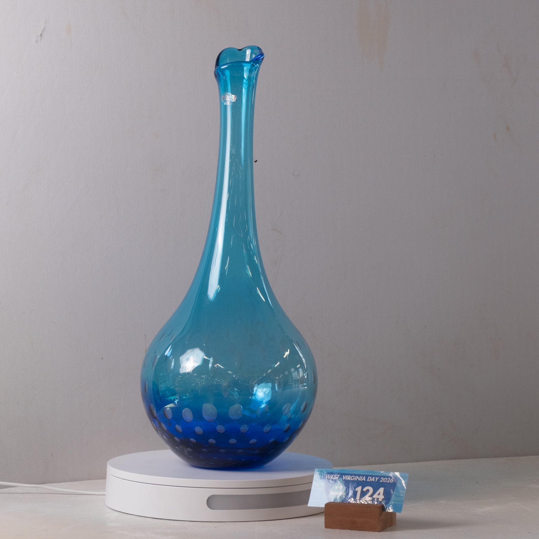 A blue glass vase with a flared neck and bubble pattern near the base, displayed on a white circular light box against a plain light gray wall.