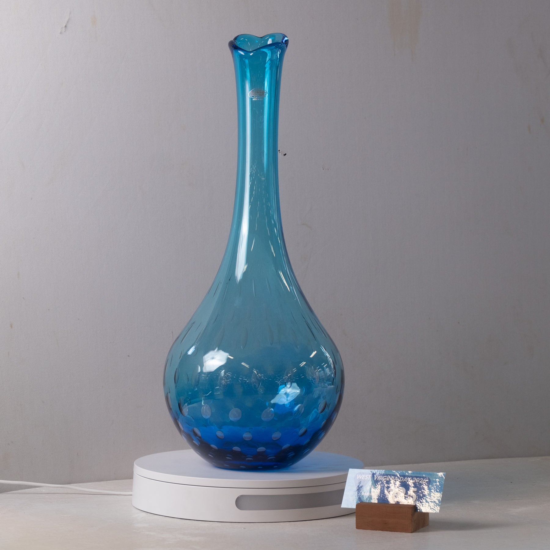 A translucent blue glass vase with a flared neck and dotted textured pattern, displayed on a white circular base against a plain light gray wall.