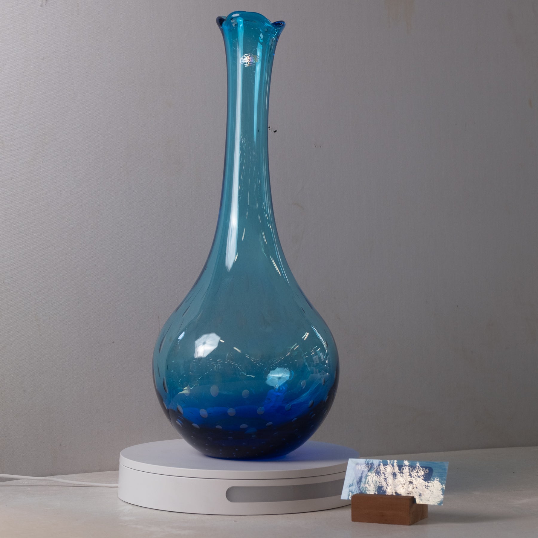 A translucent blue glass vase with a flared rim and narrow neck, displayed on a white circular base against a plain light gray wall.
