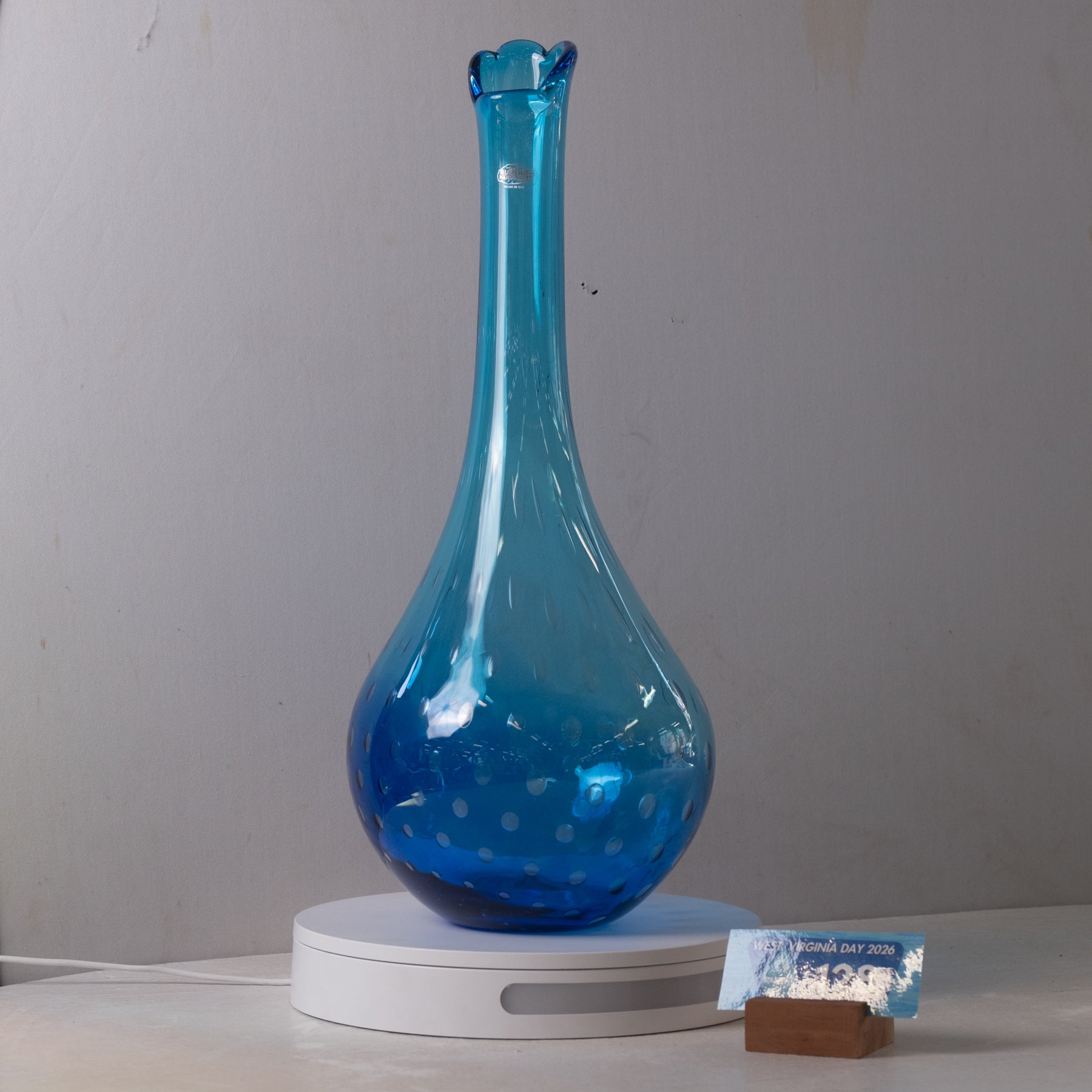 A translucent blue glass vase with a flared rim and dotted pattern near the base, displayed on a white circular stand against a plain light gray wall.
