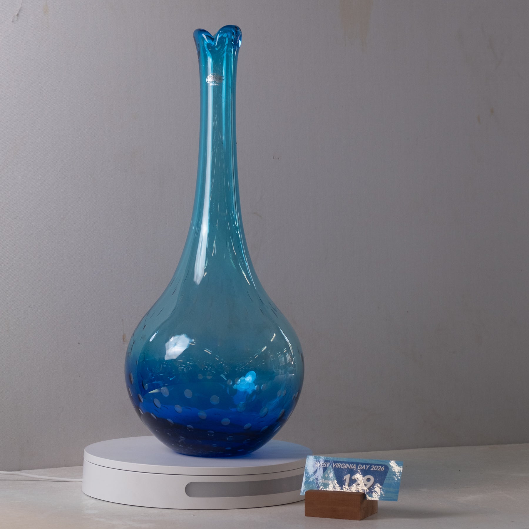 A translucent blue glass vase with a flared rim and bubble-like texture, displayed on a white circular base against a plain light gray wall.