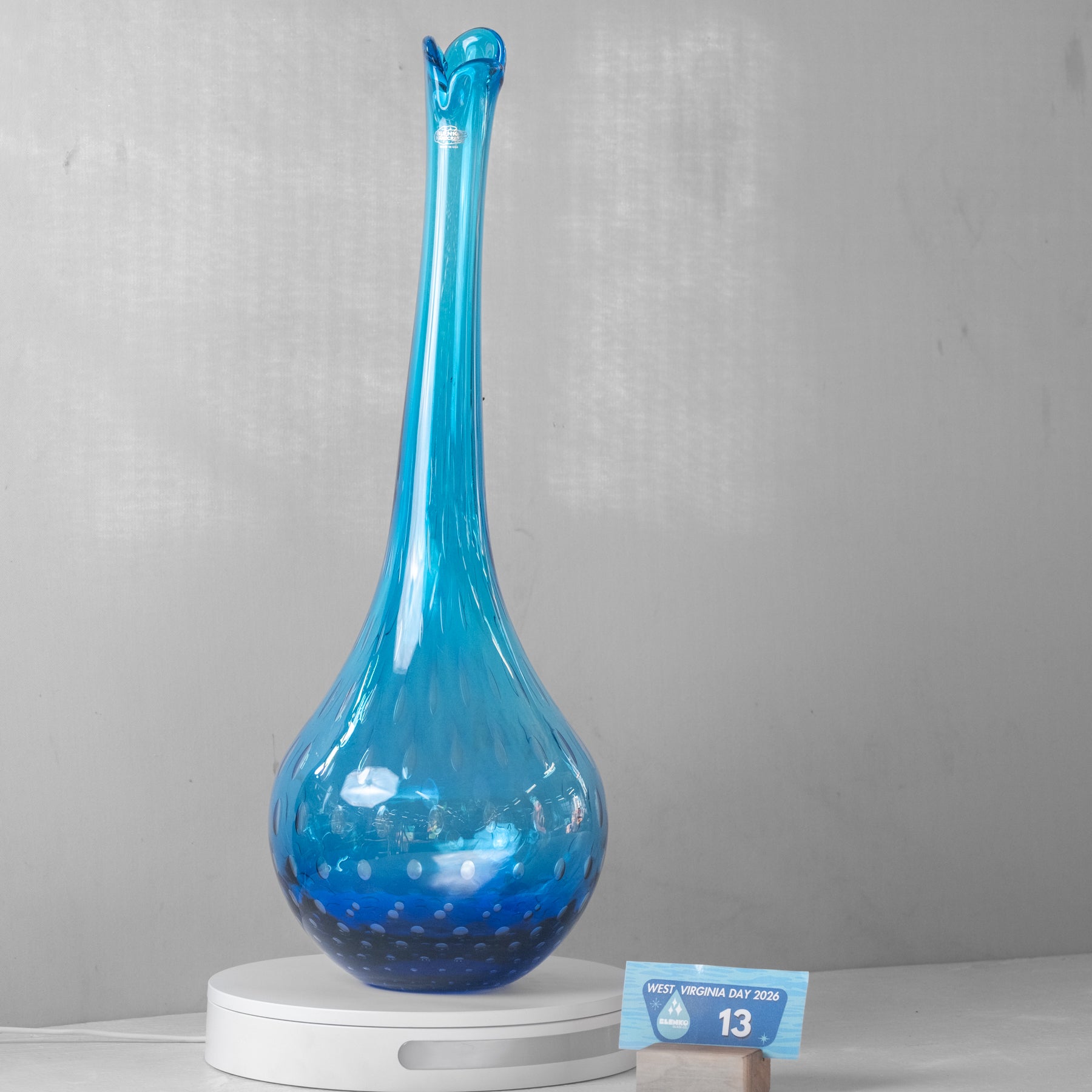 A tall, slender blue glass vase with a bulbous base and small flared rim, displayed on a white round pedestal against a light gray wall. A small blue size card is placed beside the base.