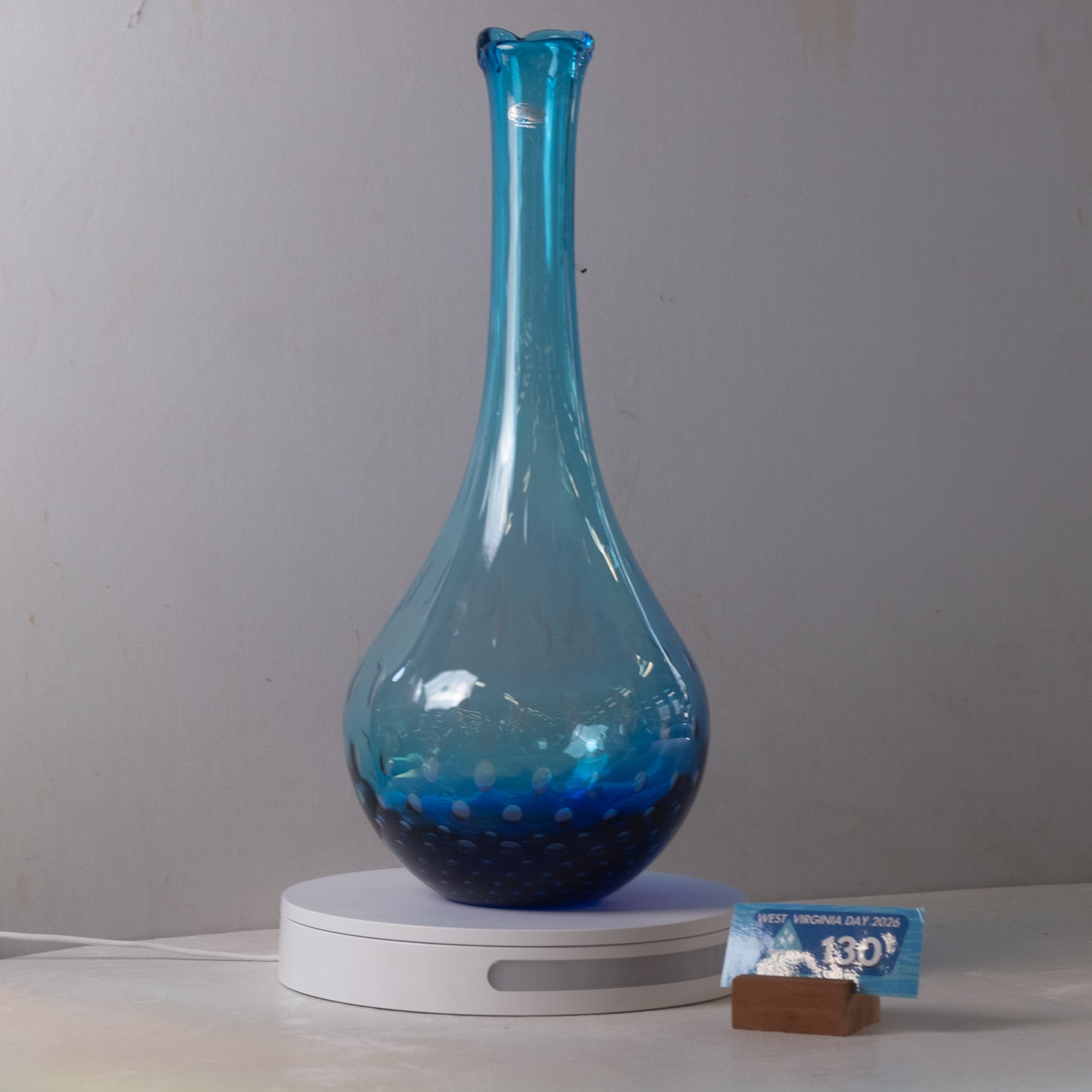 A blue glass vase with a flared neck and speckled lower section, displayed on a white circular base against a plain light gray wall.