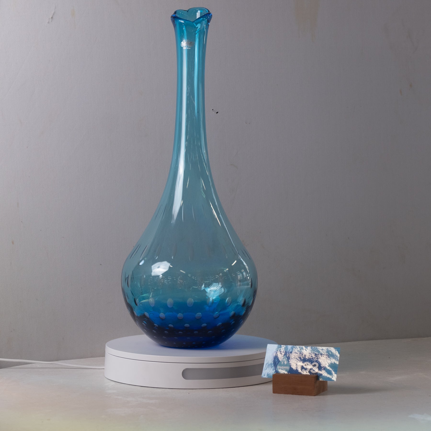 A translucent blue glass vase with a flared neck and rounded base, displayed on a white circular charging base against a plain light gray wall.