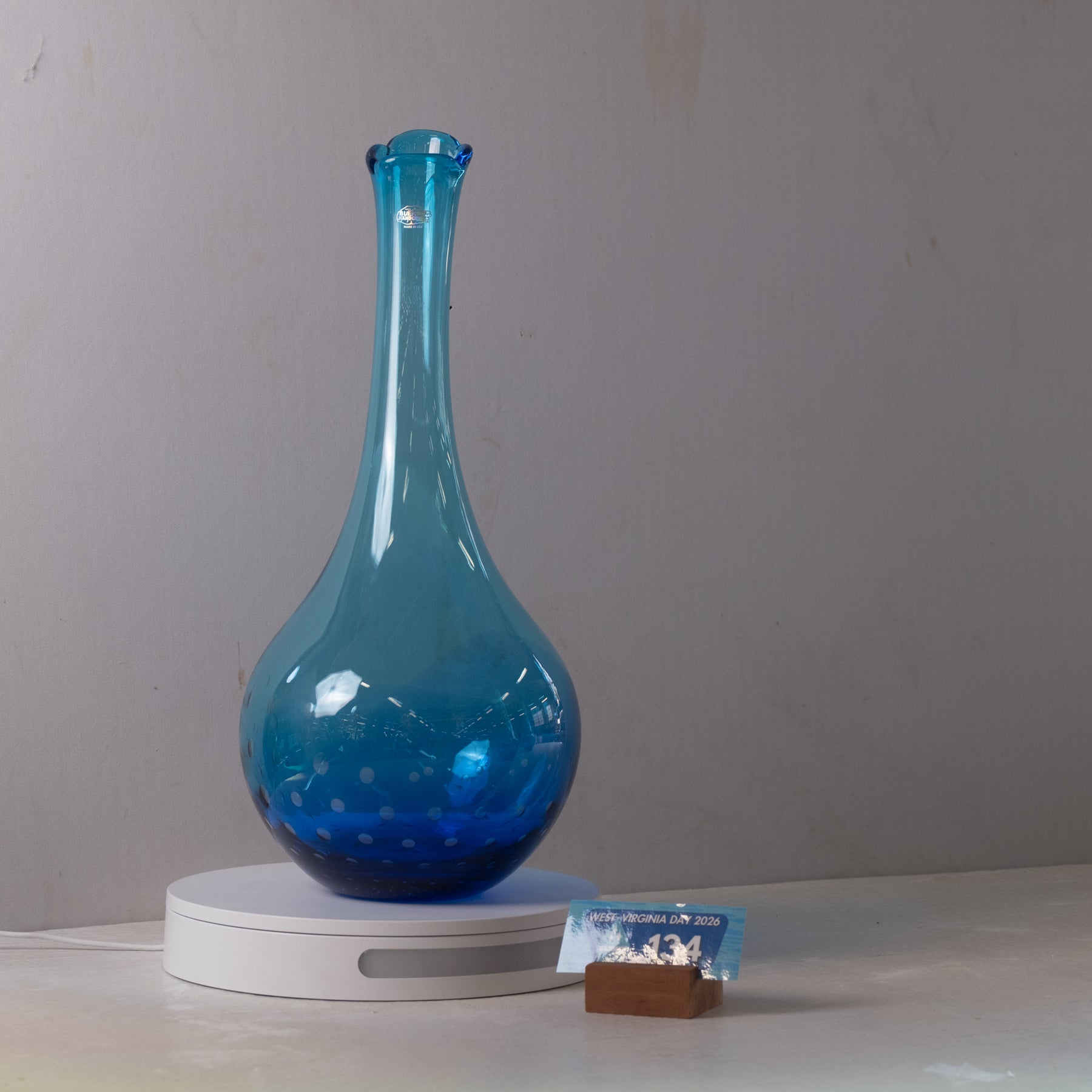 A translucent blue glass vase with a bulbous rounded body and a narrow neck, displayed on a white circular light box with a small brown block and blue packet beside it against a plain light gray wall.