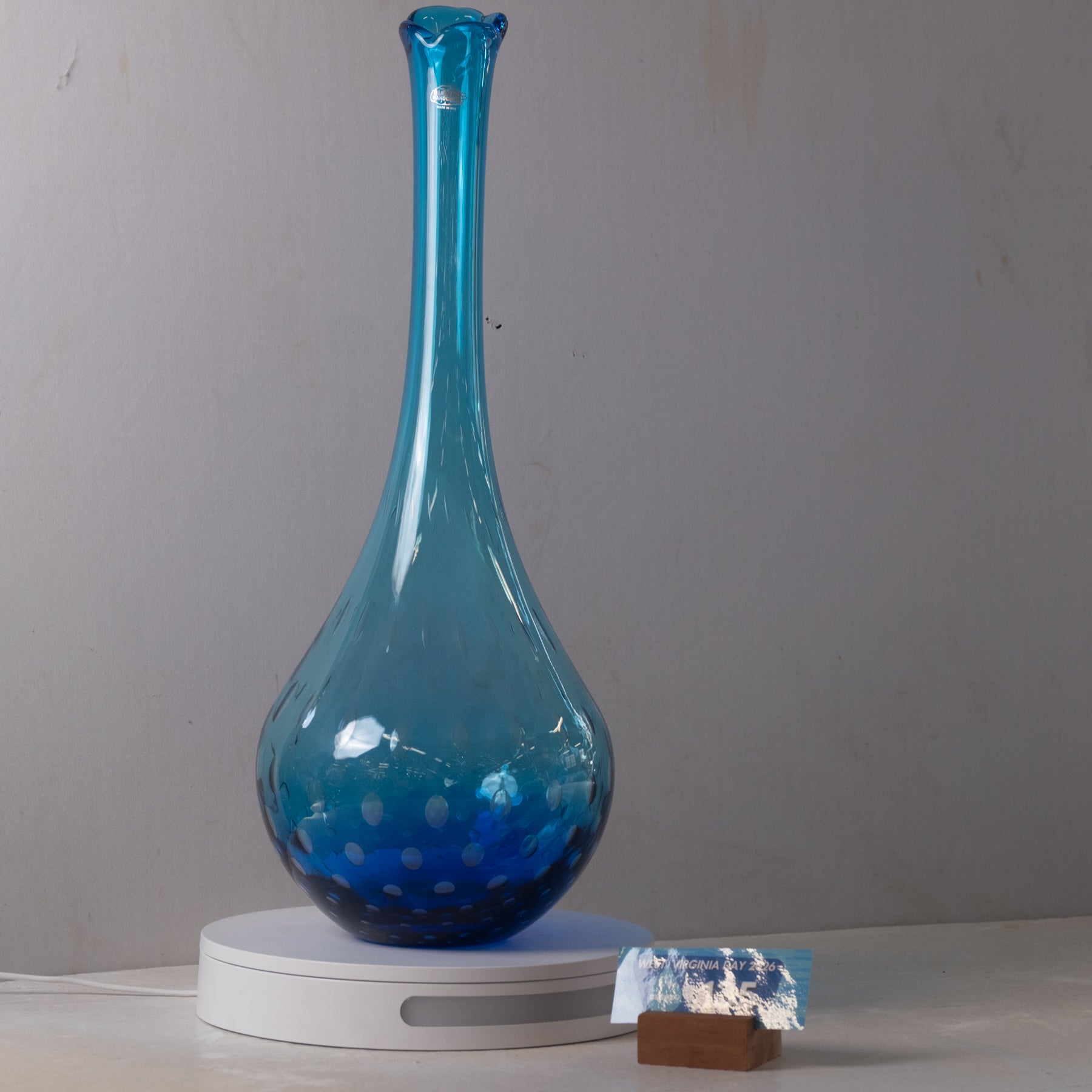 A translucent blue glass vase with a flared neck and rounded base, displayed on a white circular stand against a plain light gray wall.
