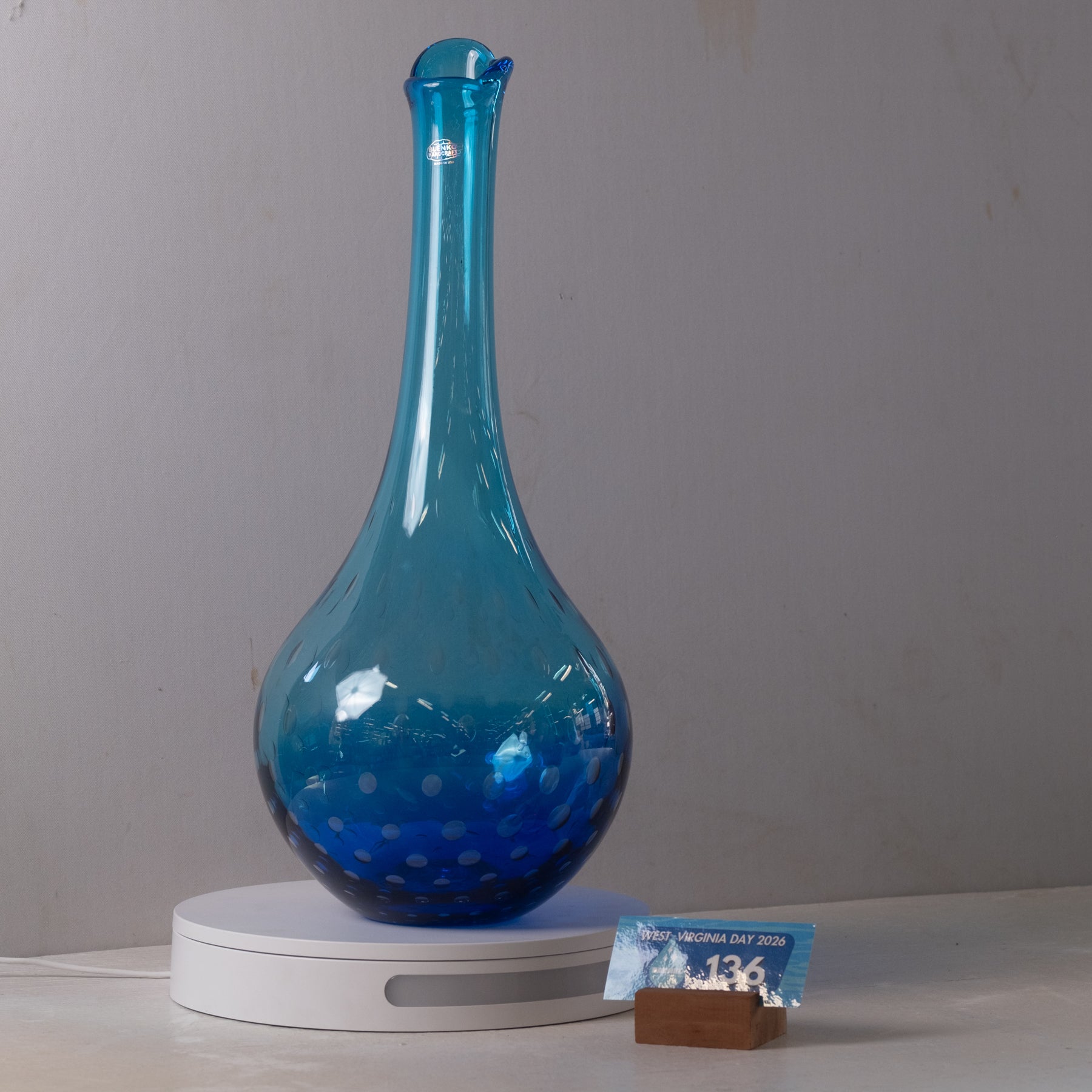 A translucent blue glass decanter with a bulbous body and narrow neck, displayed on a white circular stand against a plain light gray wall. A small printed card is placed beside the stand.