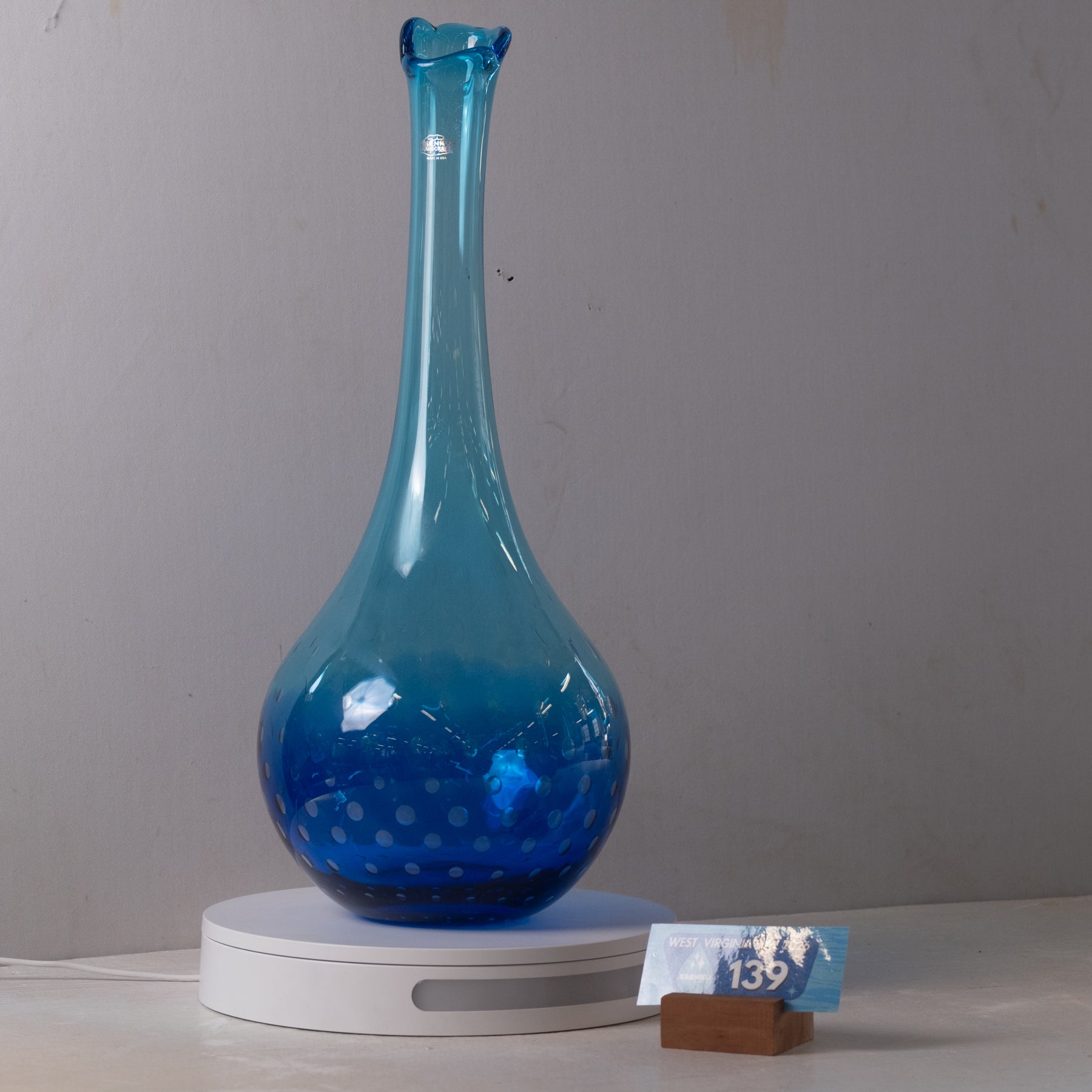 A translucent blue glass vase with a flared neck and rounded base, displayed on a white circular light box. A small price tag reading 139 is visible in front on a tabletop against a plain gray wall.