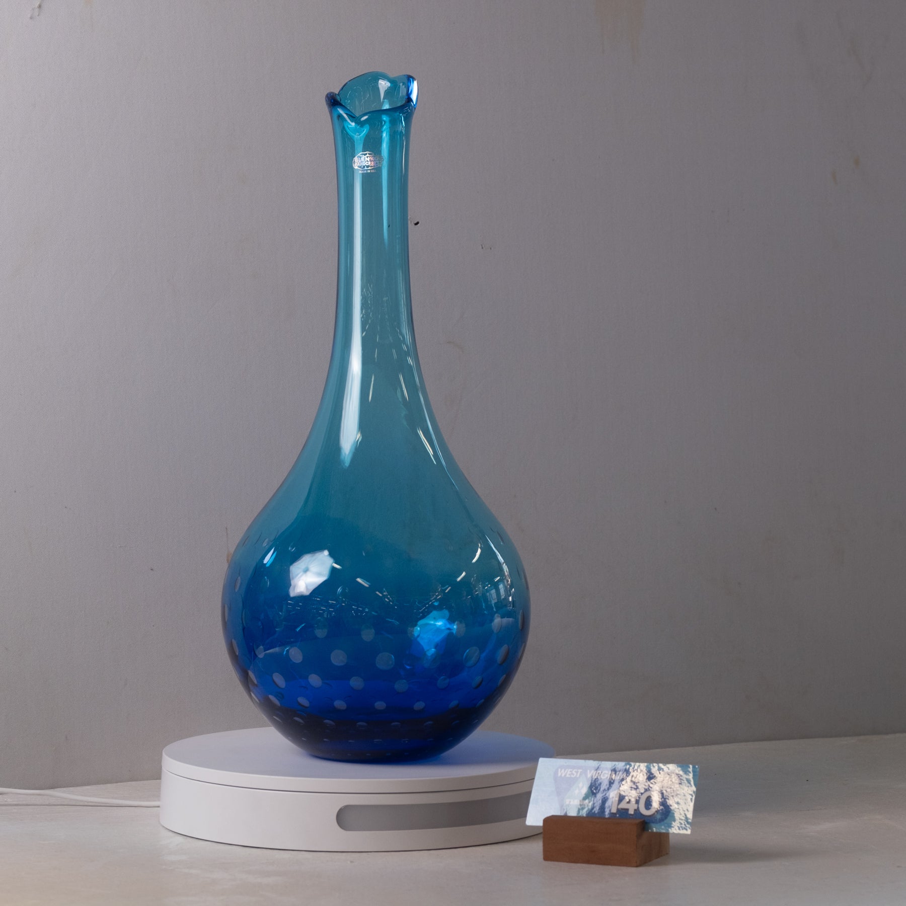 A translucent blue glass vase with a bulbous body and narrow flared neck, displayed on a white circular stand against a plain light gray wall.