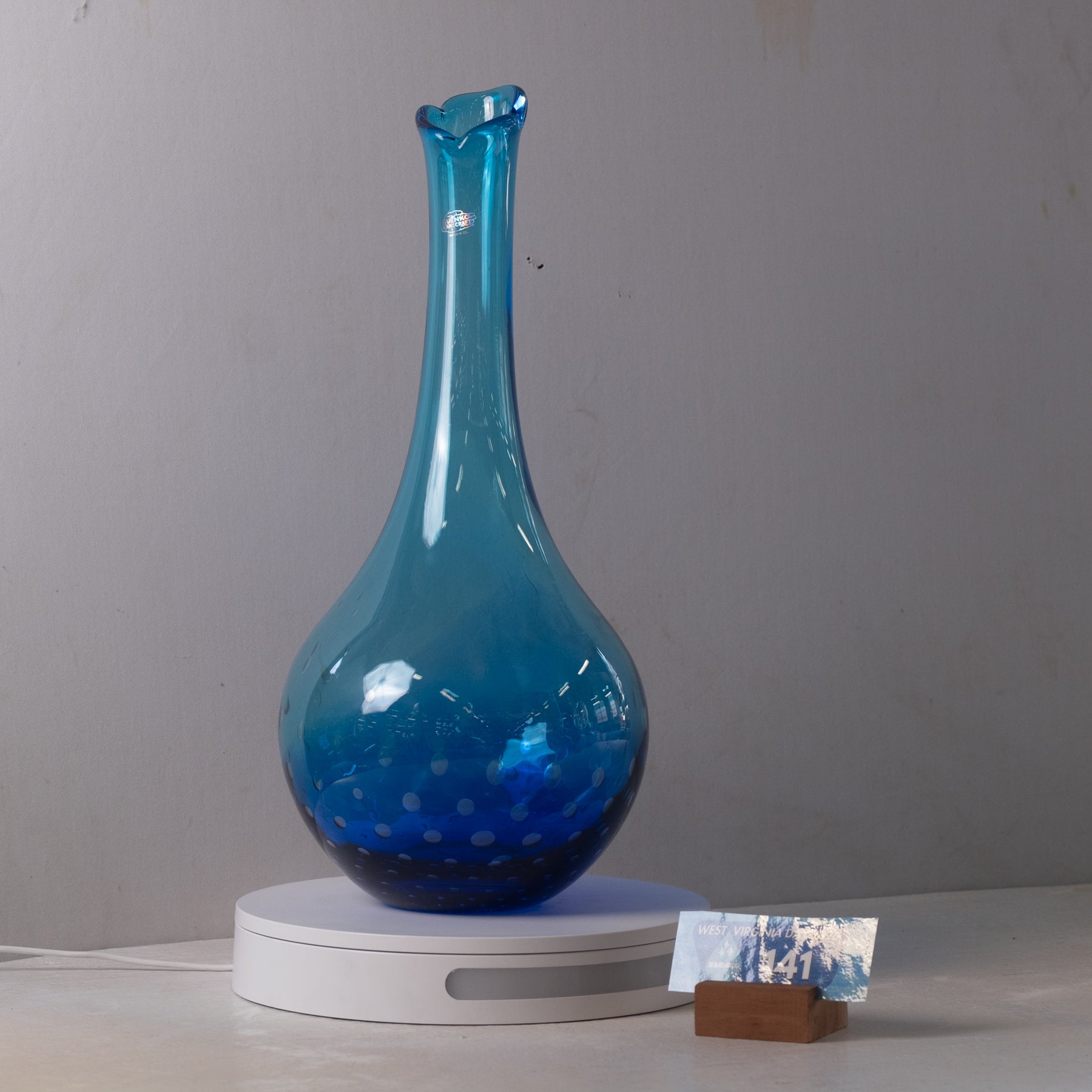 A blue glass vase with a flared neck and subtle dotted pattern near the base, displayed on a white circular stand against a plain gray wall.