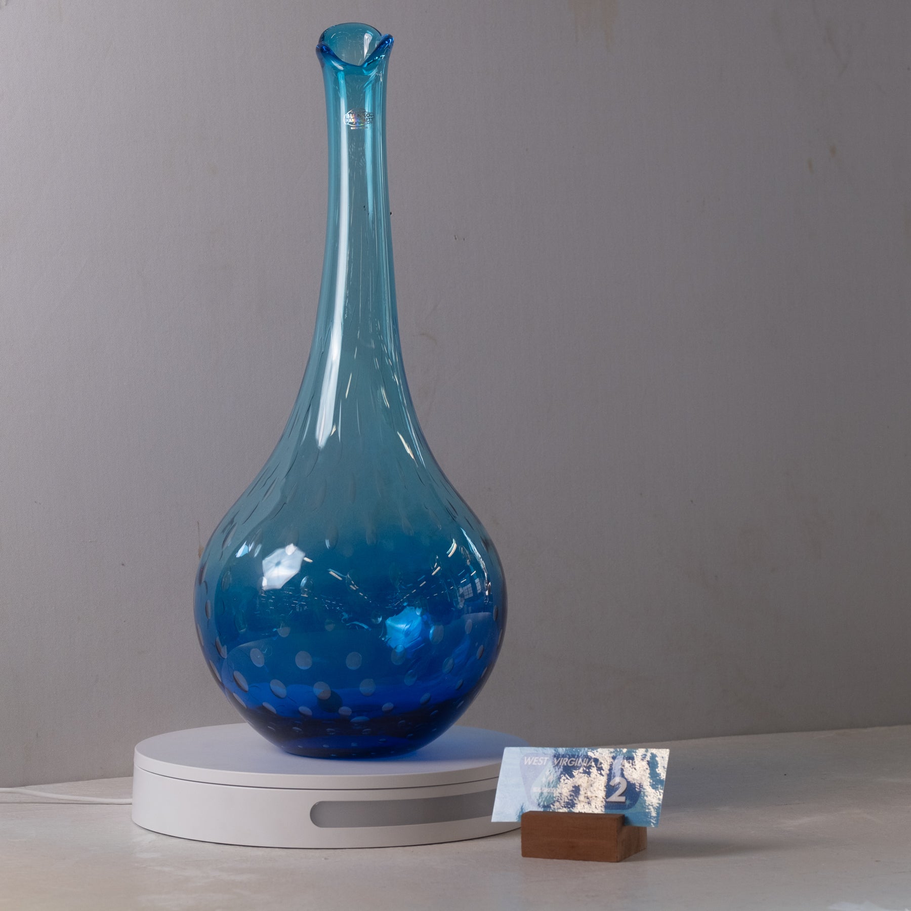 A translucent blue glass vase with a bulbous base and narrow neck, displayed on a white circular stand against a plain light gray wall.