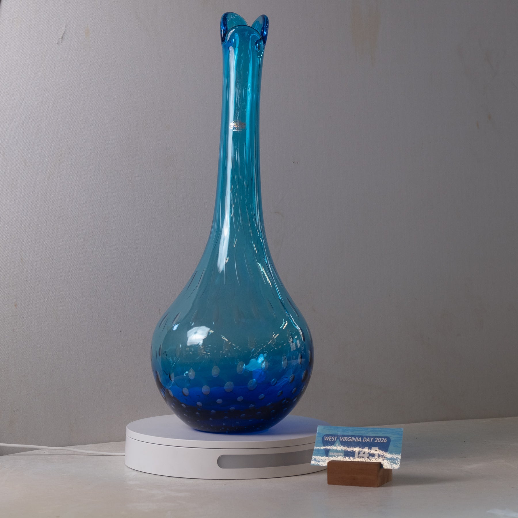 A blue glass vase with a long neck and flared rim, displayed on a white circular stand against a plain light gray wall. A small card and brown block are placed beside the stand.