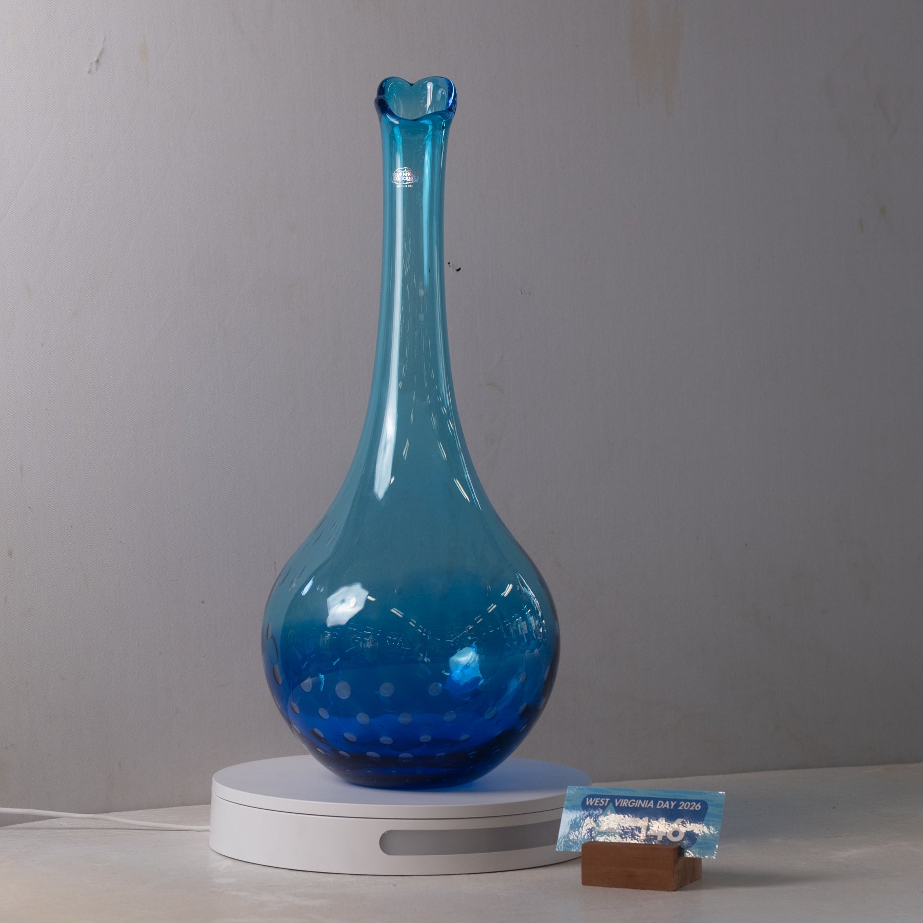 A translucent blue glass vase with a flared rim and bulbous base, displayed on a white circular light box against a plain light gray wall.