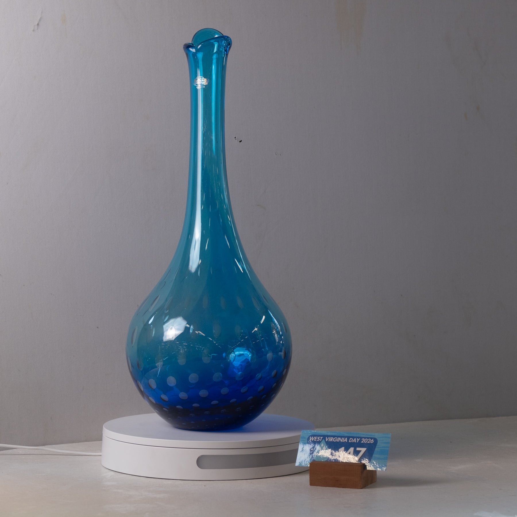 A translucent cobalt blue glass vase with a flared neck and subtle bubble pattern near the base, displayed on a white circular light box. A small card and brown block are placed beside the base against a plain gray wall.