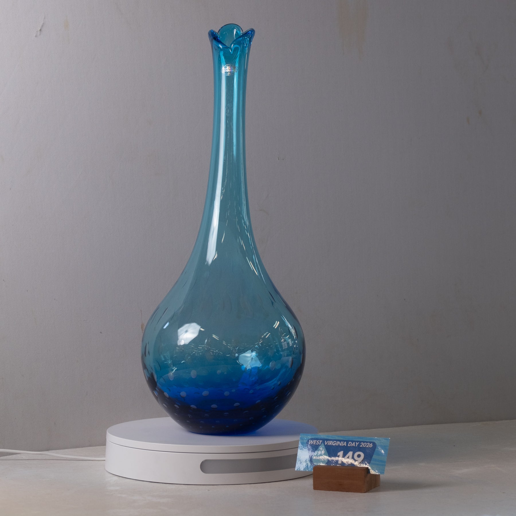 A translucent blue glass vase with a flared rim and bulbous base, displayed on a white round pedestal against a plain light gray wall. A small price tag card is placed beside the pedestal.