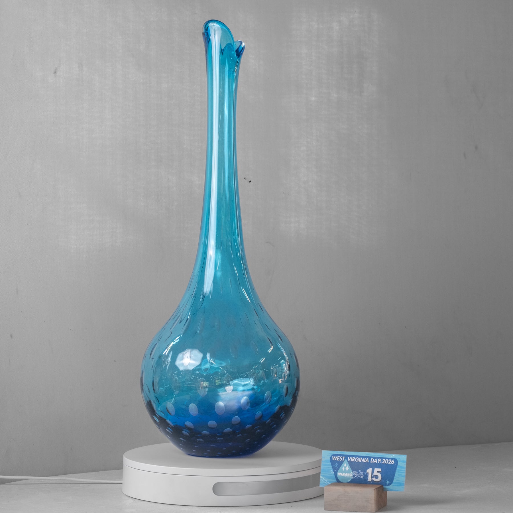 A tall, slender blue glass vase with a bulbous base and a narrow flared neck, displayed on a white circular charging base against a gray wall. A small Arizona state flag card is placed beside the base.