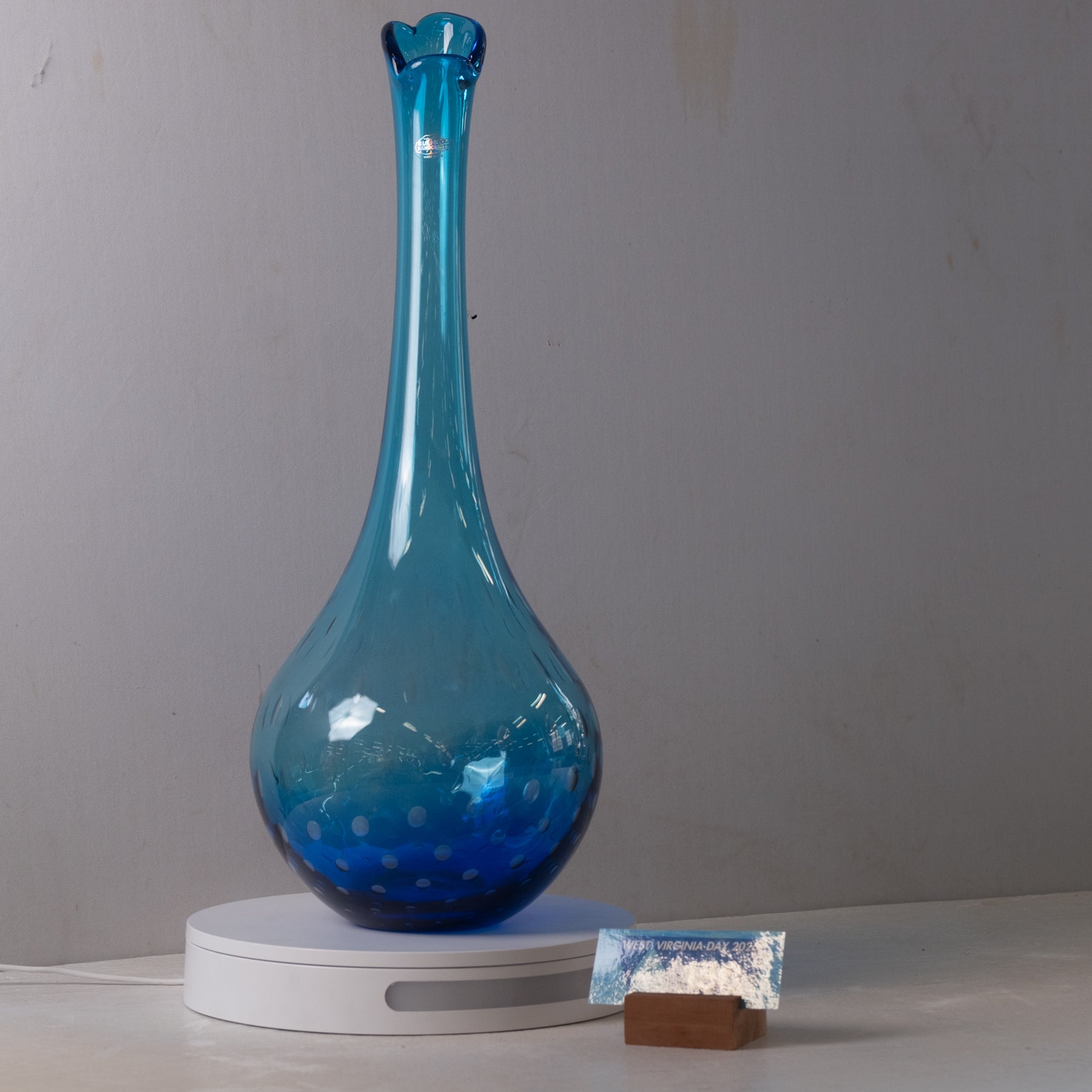 A tall, slender blue glass vase with a flared rim and subtle white speckled pattern near the base, displayed on a white circular scale against a plain light gray wall.