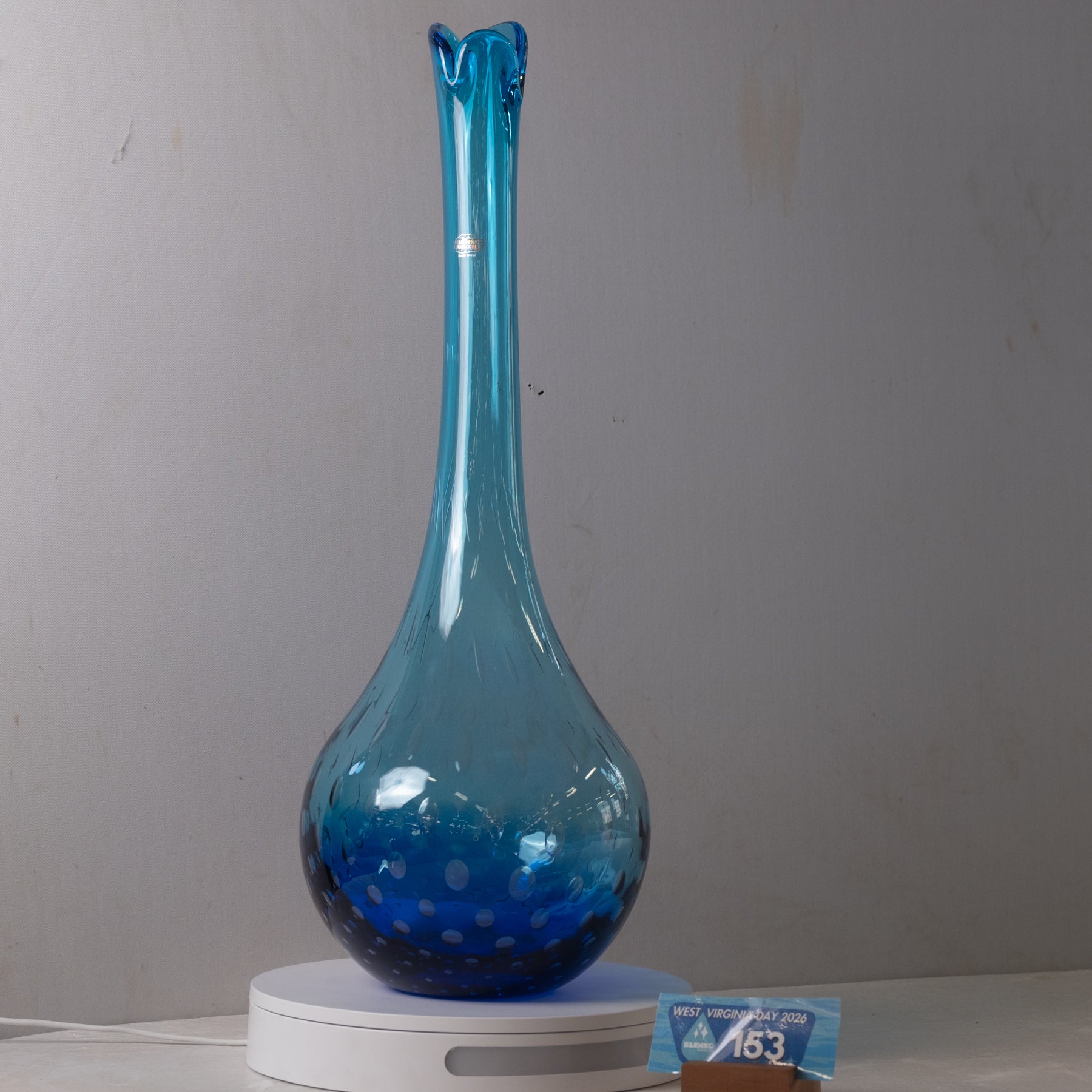 A tall, narrow blue glass vase with a flared rim and a bulbous base, displayed on a white circular light box against a plain gray wall.