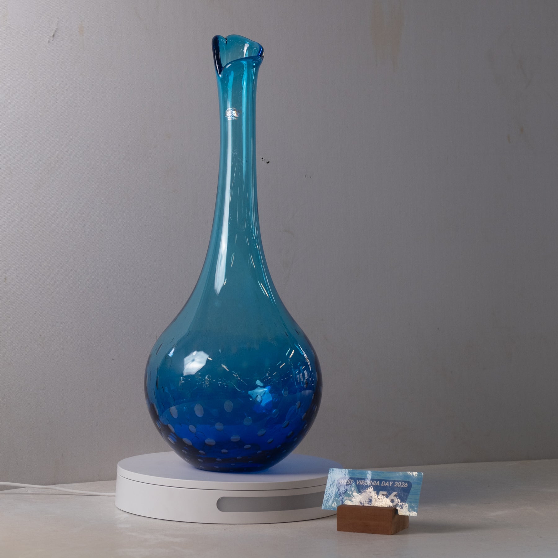 A translucent blue glass decanter with a flared neck and small spout, resting on a white circular display base against a plain gray wall.