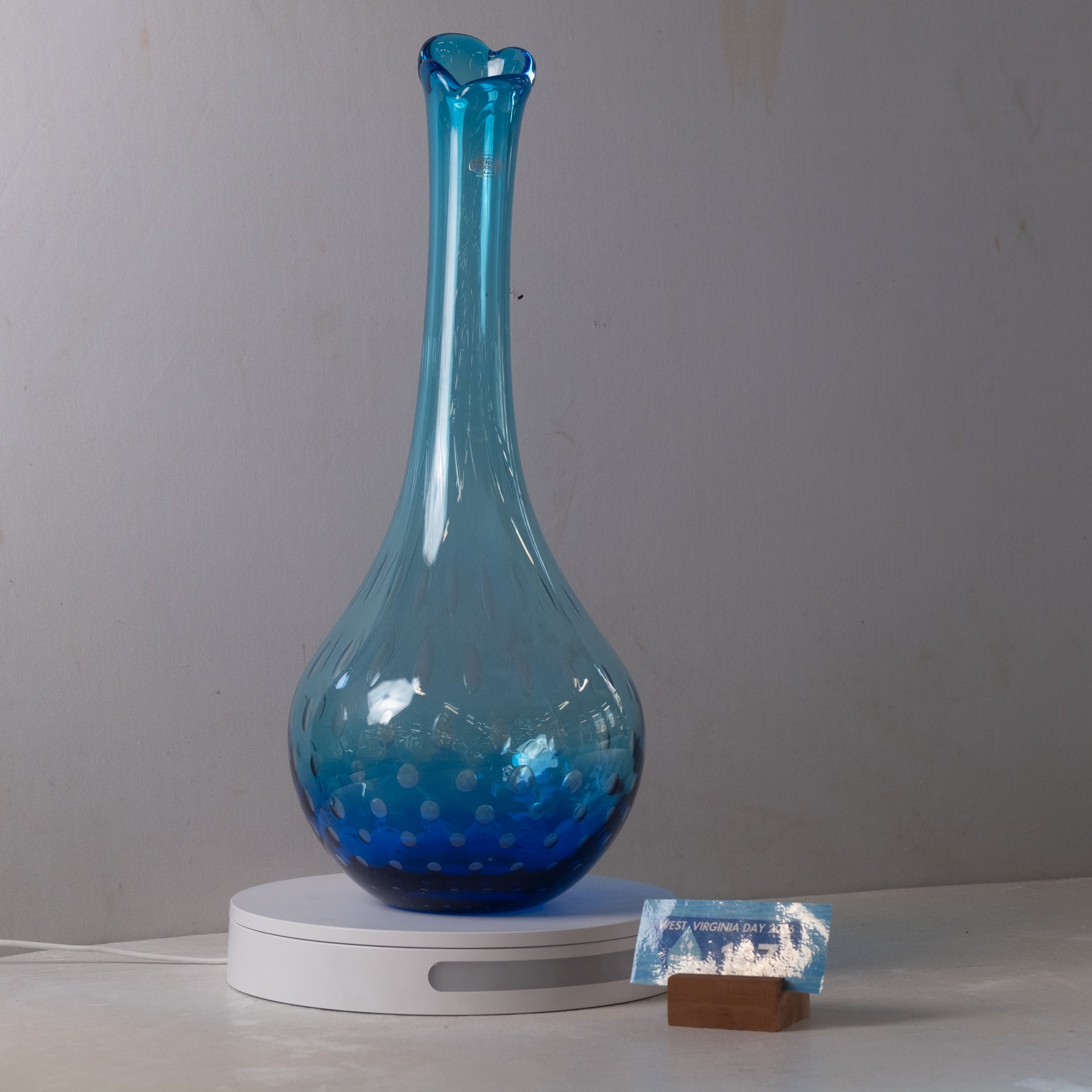 A translucent blue glass vase with a flared neck and dotted pattern near the base, displayed on a white circular light box against a plain gray wall.