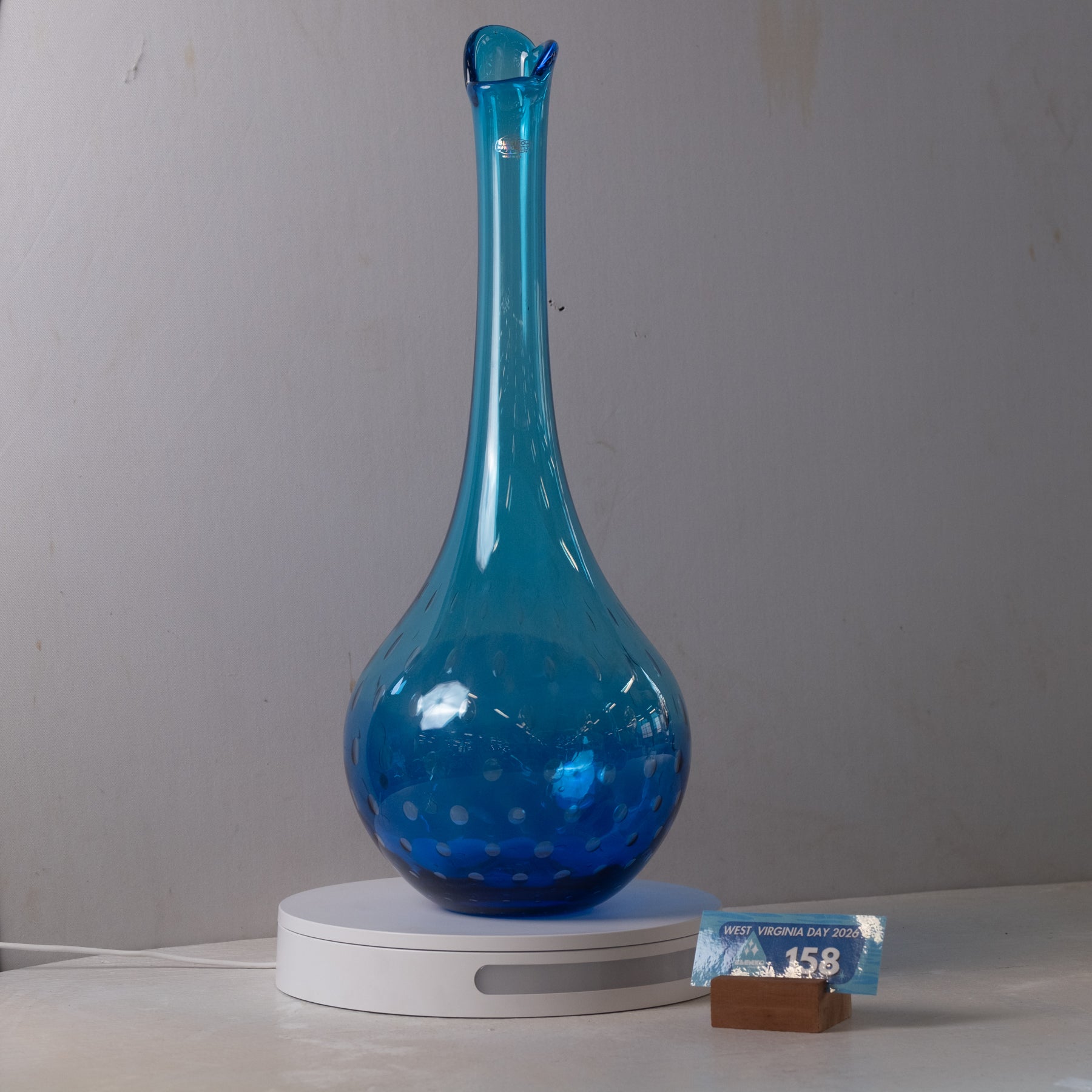 A translucent cobalt blue glass decanter with a long neck and small spout, displayed on a white digital scale with a small price tag in the foreground against a plain gray wall.