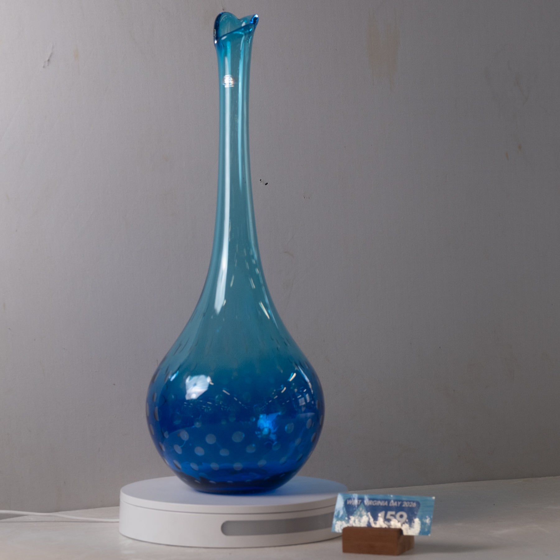 A blue glass decanter with a flared spout and dotted pattern, displayed on a white circular base against a plain gray wall.
