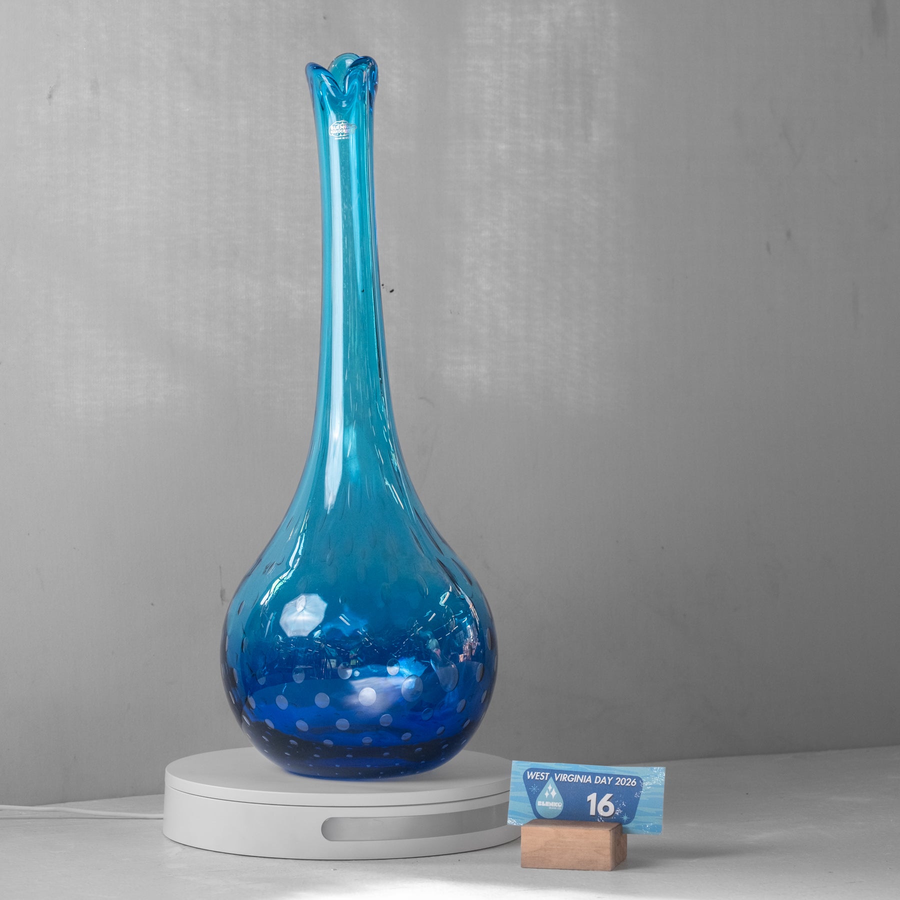 A tall, bulb-shaped blue glass vase with a flared neck and small circular dot pattern near the base, displayed on a white circular pedestal. A small sign stands in front of the pedestal against a plain gray wall.