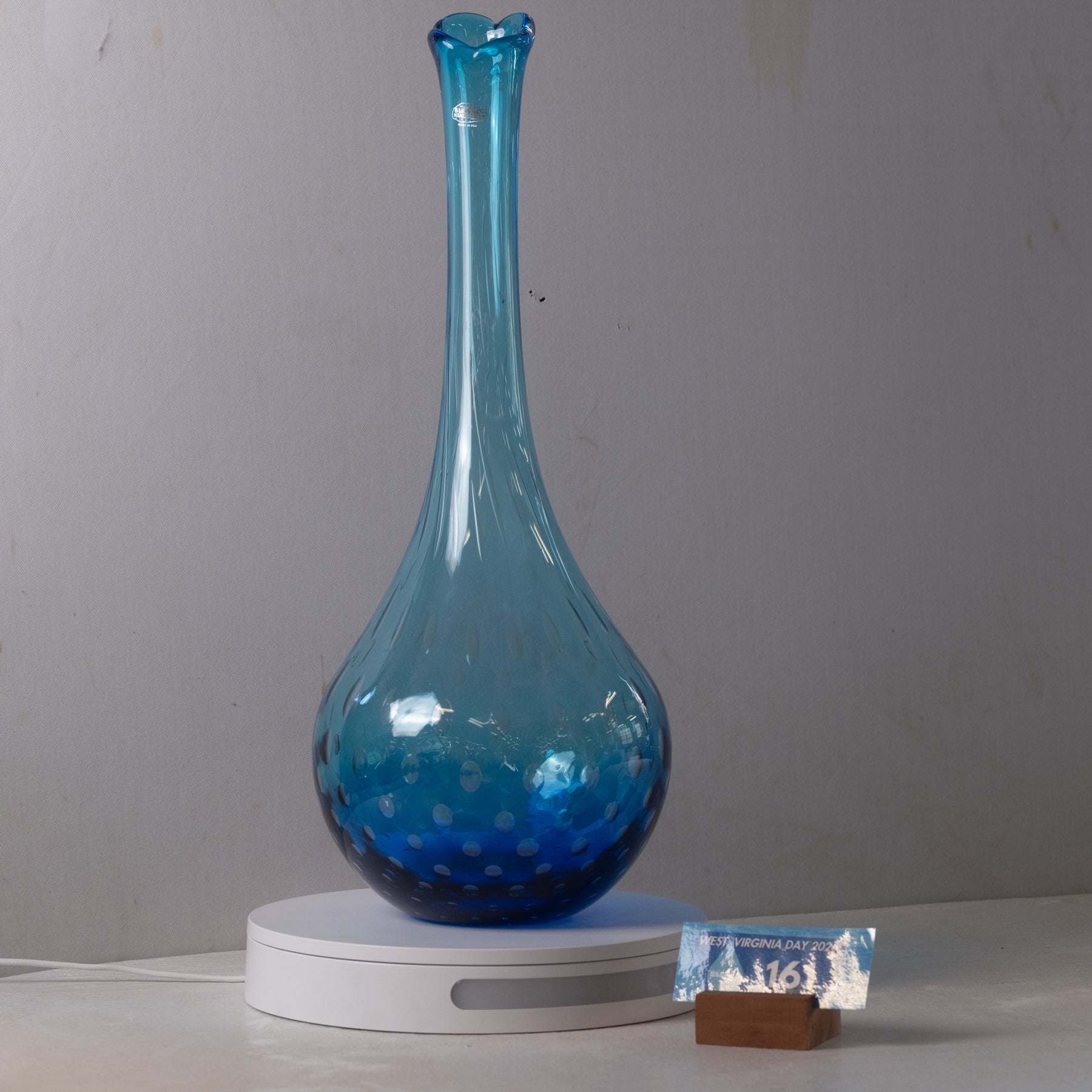 A tall, bulb-shaped blue glass vase with a flared neck and small white speckles near the base, displayed on a white circular light box with a small branded card on a wooden block beside it.