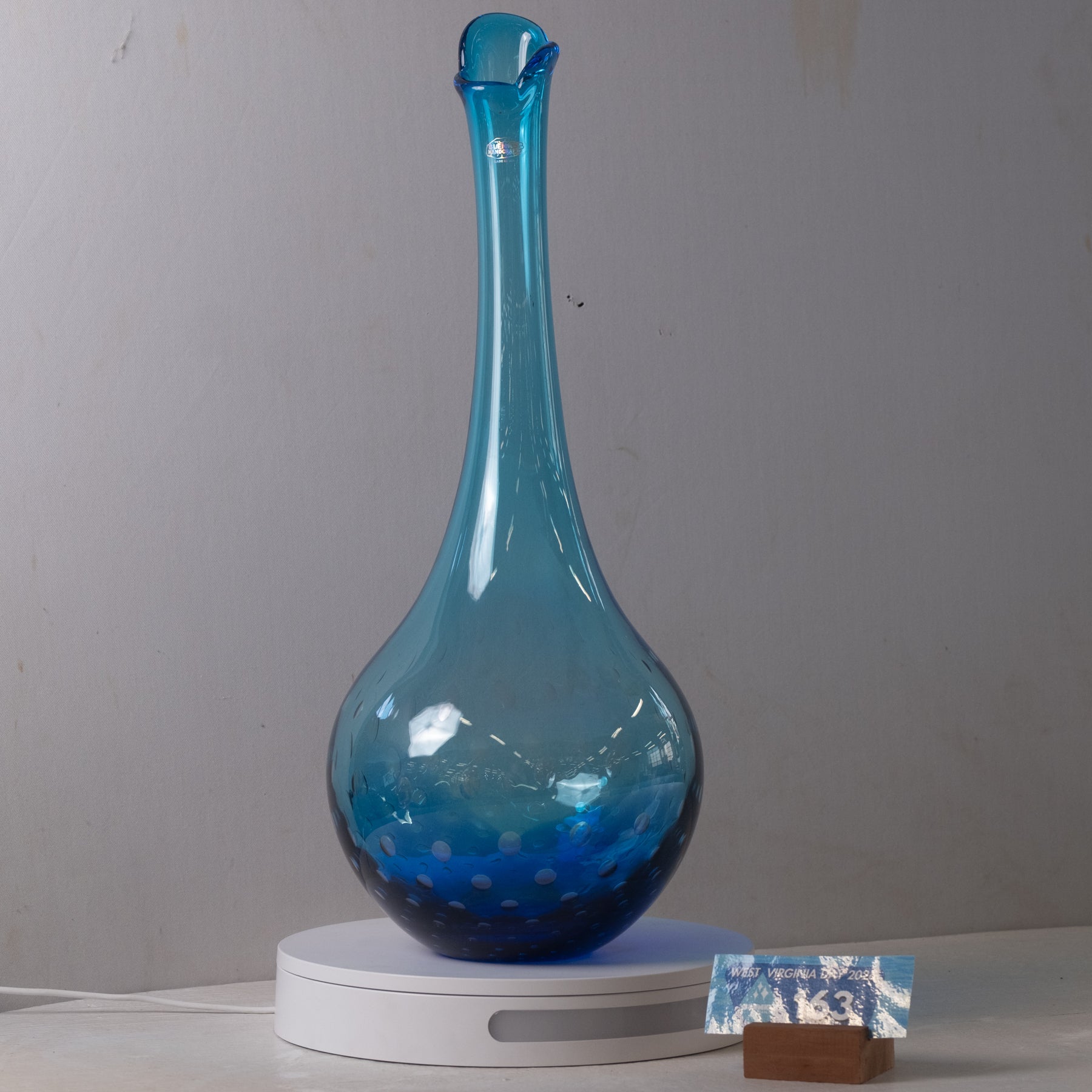 A blue glass decanter with a long neck and small spout, displayed on a white circular charging base against a plain gray wall.