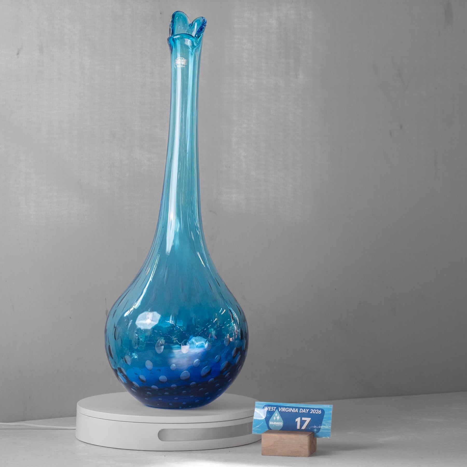 A tall, slender blue glass vase with a flared rim and bubble-like speckled texture, displayed on a white round pedestal against a gray wall. A small blue price card on a wooden block sits beside the pedestal.