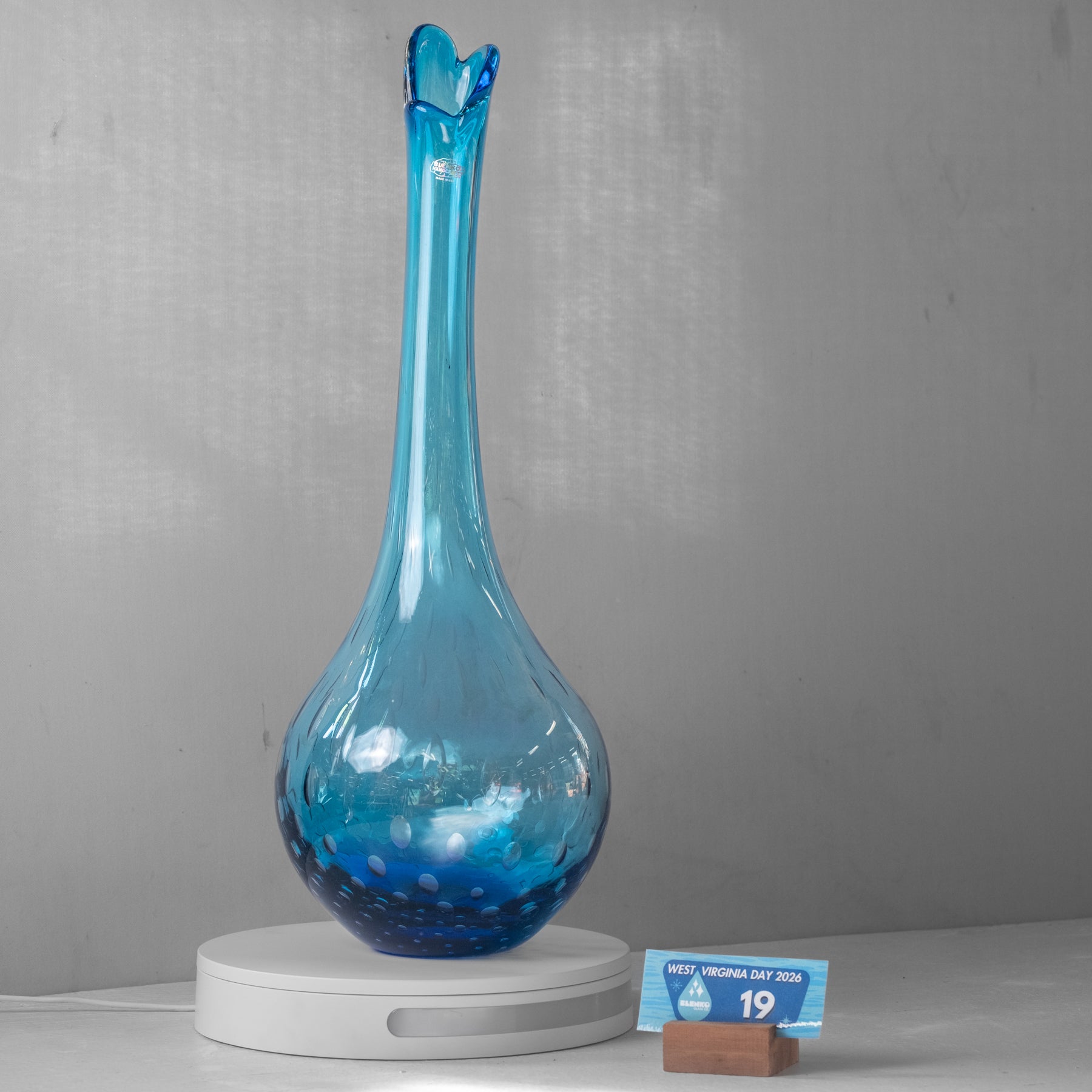 A tall, bulb-shaped blue glass vase with a flared, ruffled neck sits on a white circular charging base against a gray wall. A small card on a wooden stand is placed beside the base.
