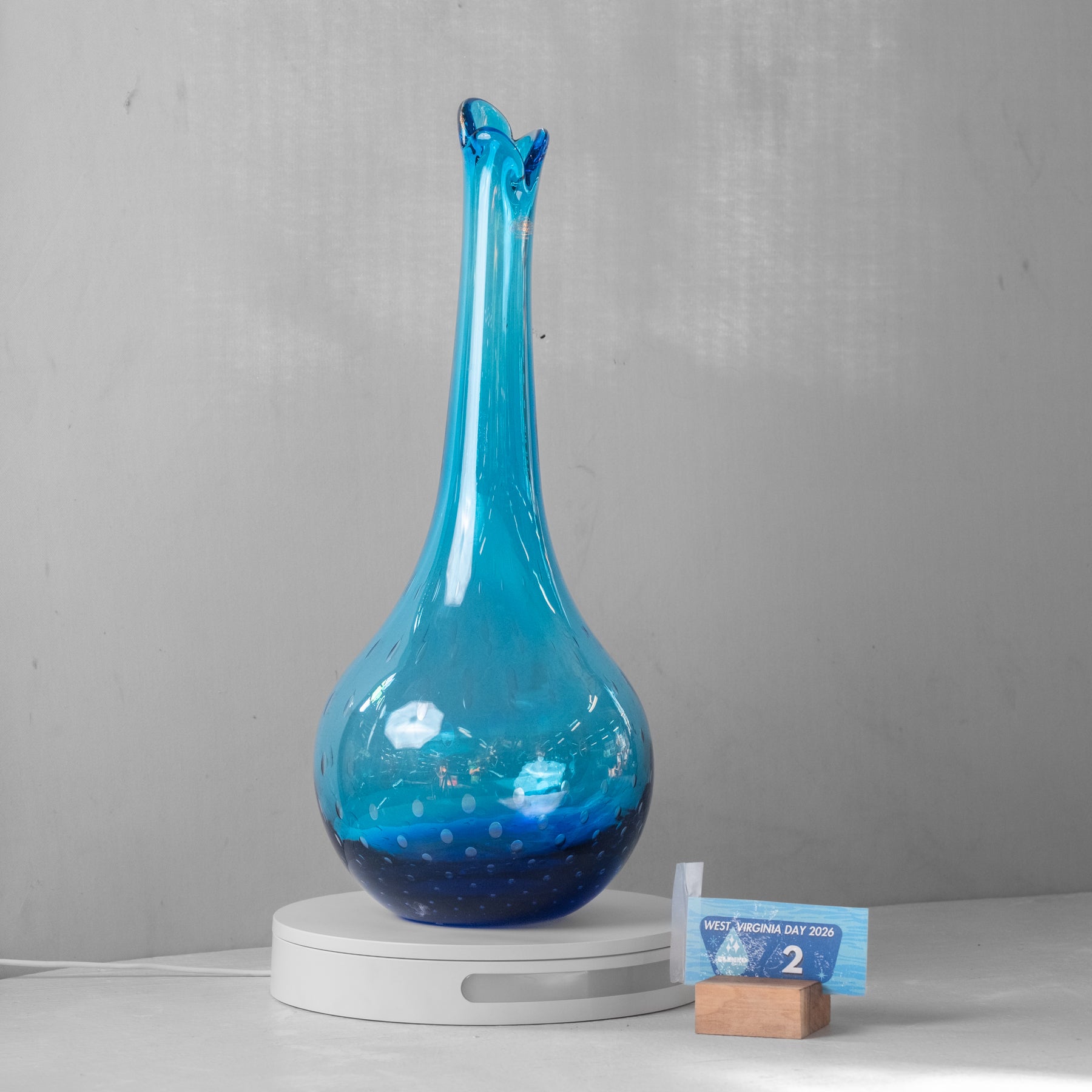 A blue glass vase with a long, narrow neck and a bulbous rounded base, displayed on a white circular pedestal. A small blue size card on a wooden block is placed beside it against a light gray wall background.