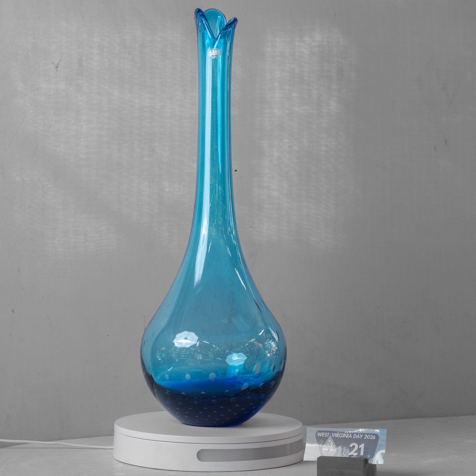 A tall, slender blue glass vase with a flared rim sits on a white circular base against a gray wall. A small card is visible at the lower right.
