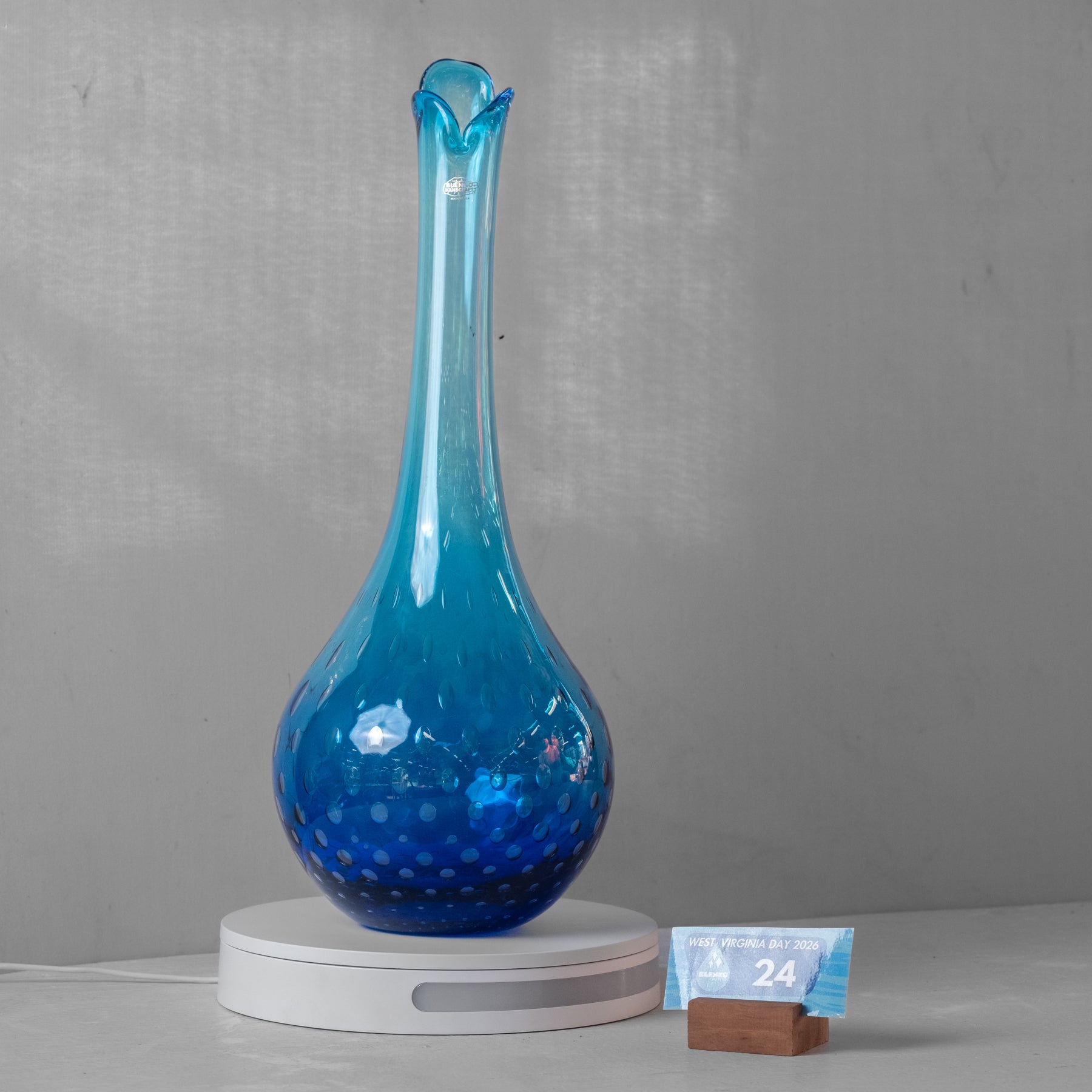 A blue glass decanter with a flared rim and dotted textured pattern, resting on a white circular scale with a small weight card next to it, set against a gray wall.