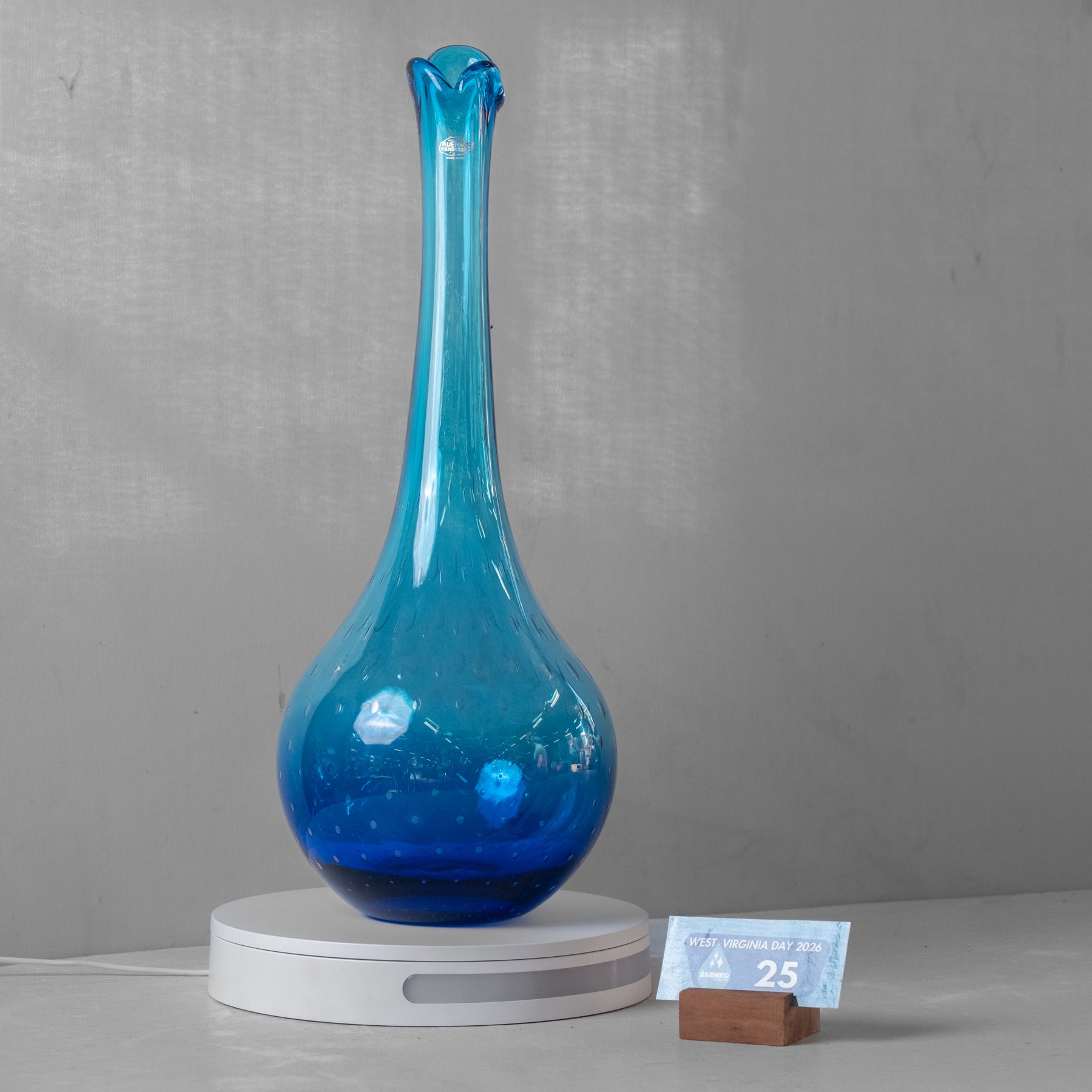 A tall, slender cobalt blue glass vase with a bulbous base, displayed on a white circular scale with a small card on a wooden block next to it, against a textured gray background.