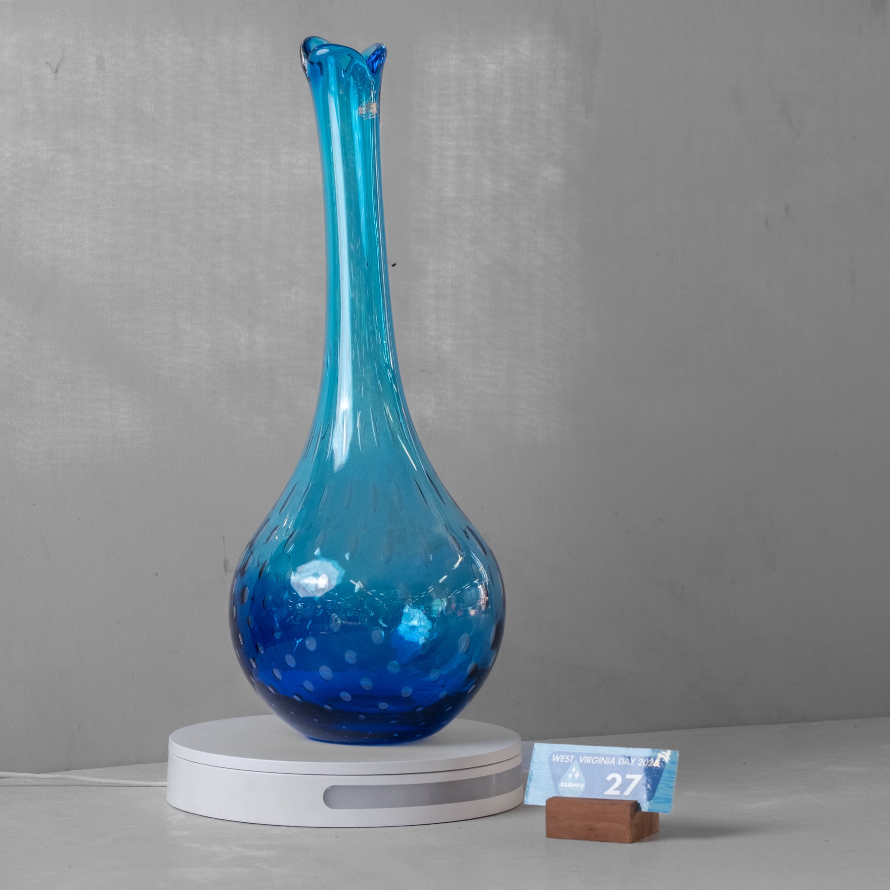 A blue glass vase with a flared neck and speckled finish, displayed on a white circular base against a gray wall.