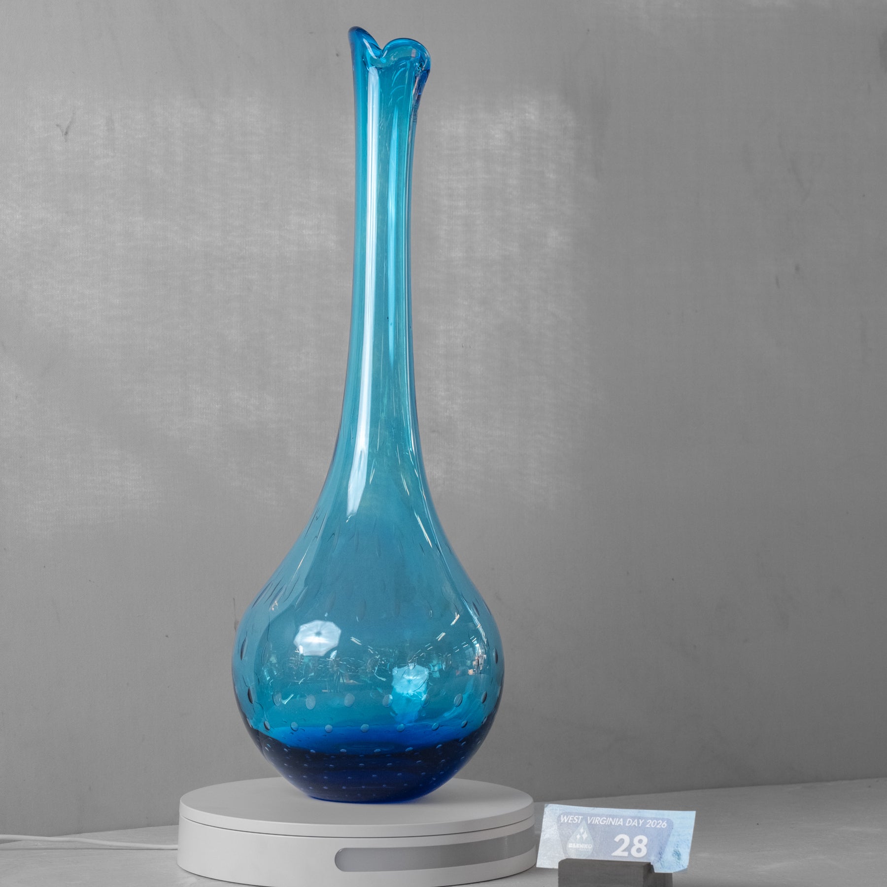 A tall, slender blue glass vase with a flared neck and bulbous base, displayed on a white round pedestal against a gray wall. A small price tag is visible at the lower right.