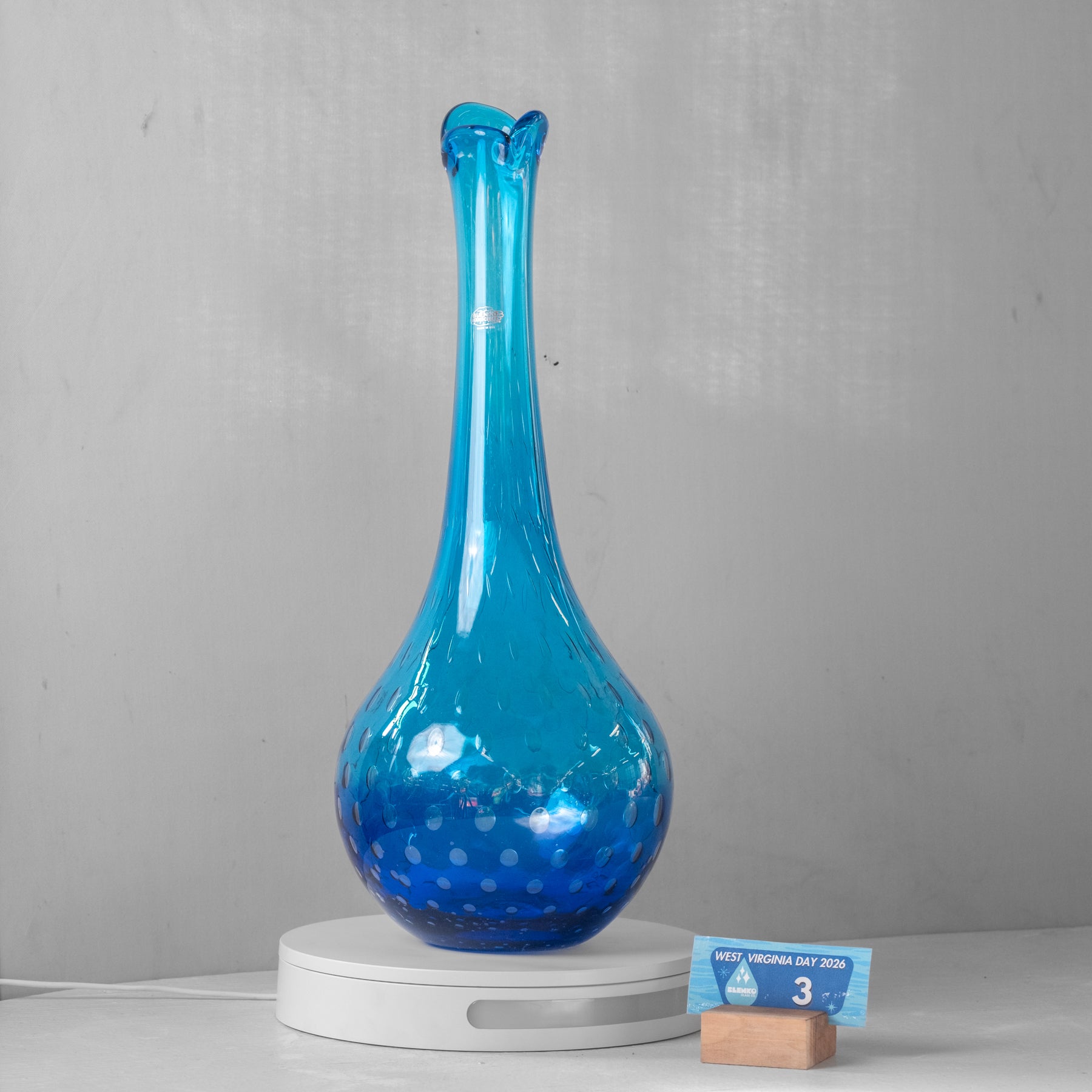 A translucent blue glass vase with a flared neck and bubble-patterned base, displayed on a white circular stand against a light gray wall.