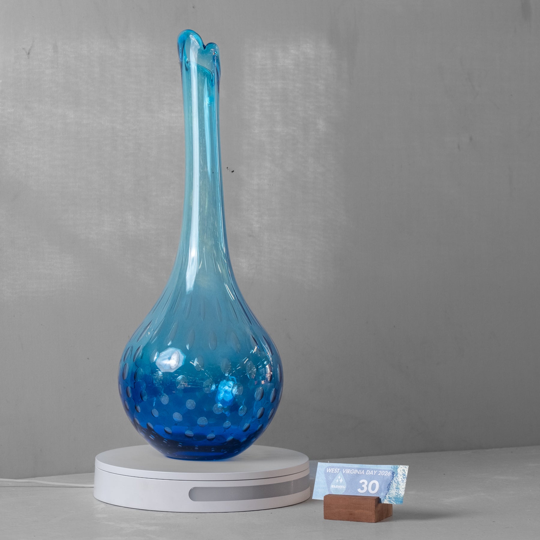 A blue glass vase with a bulbous base and a long, narrow neck, displayed on a white circular lamp base against a gray wall.
