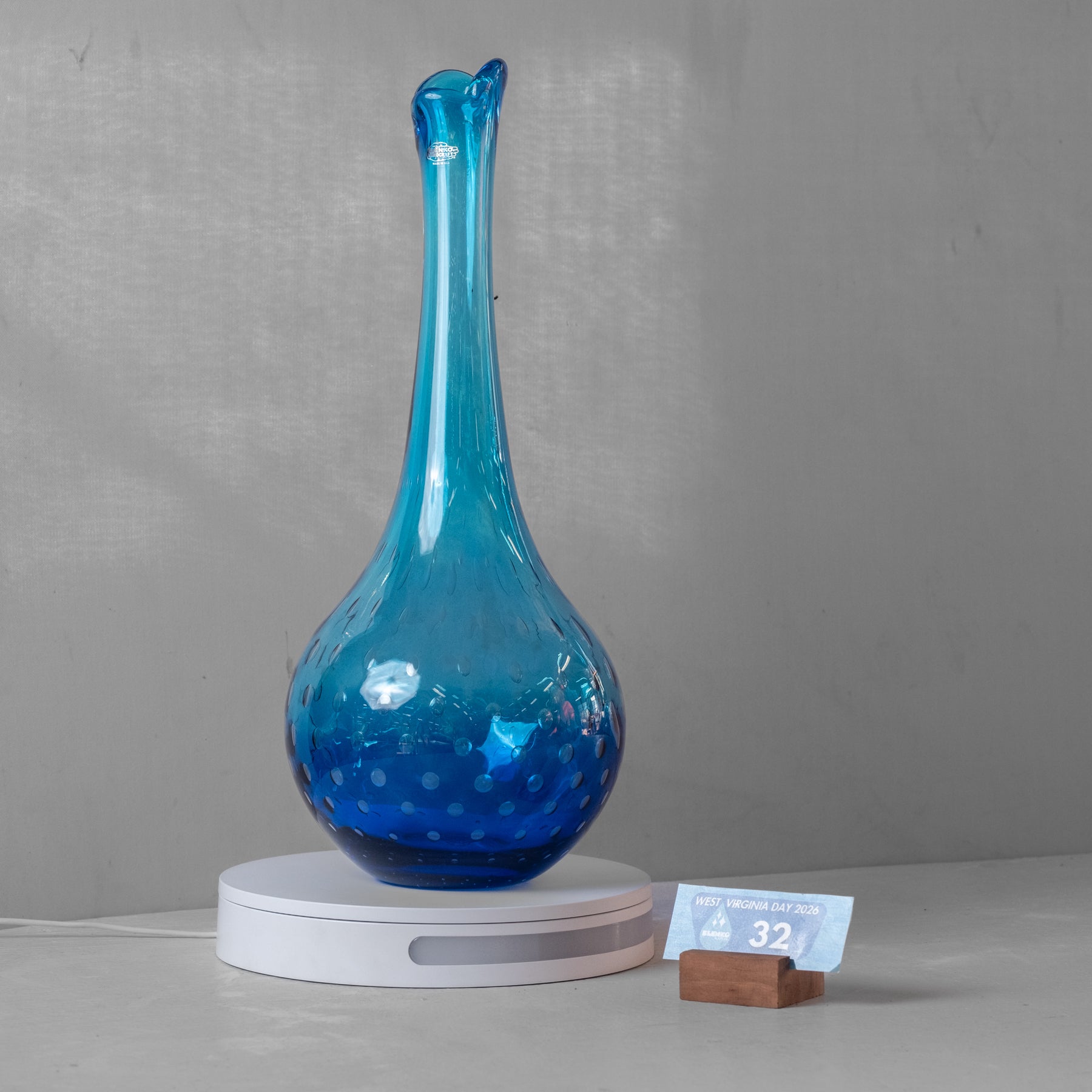 A blue glass decanter with a bulbous body and narrow neck, displayed on a white circular charging base against a gray background.