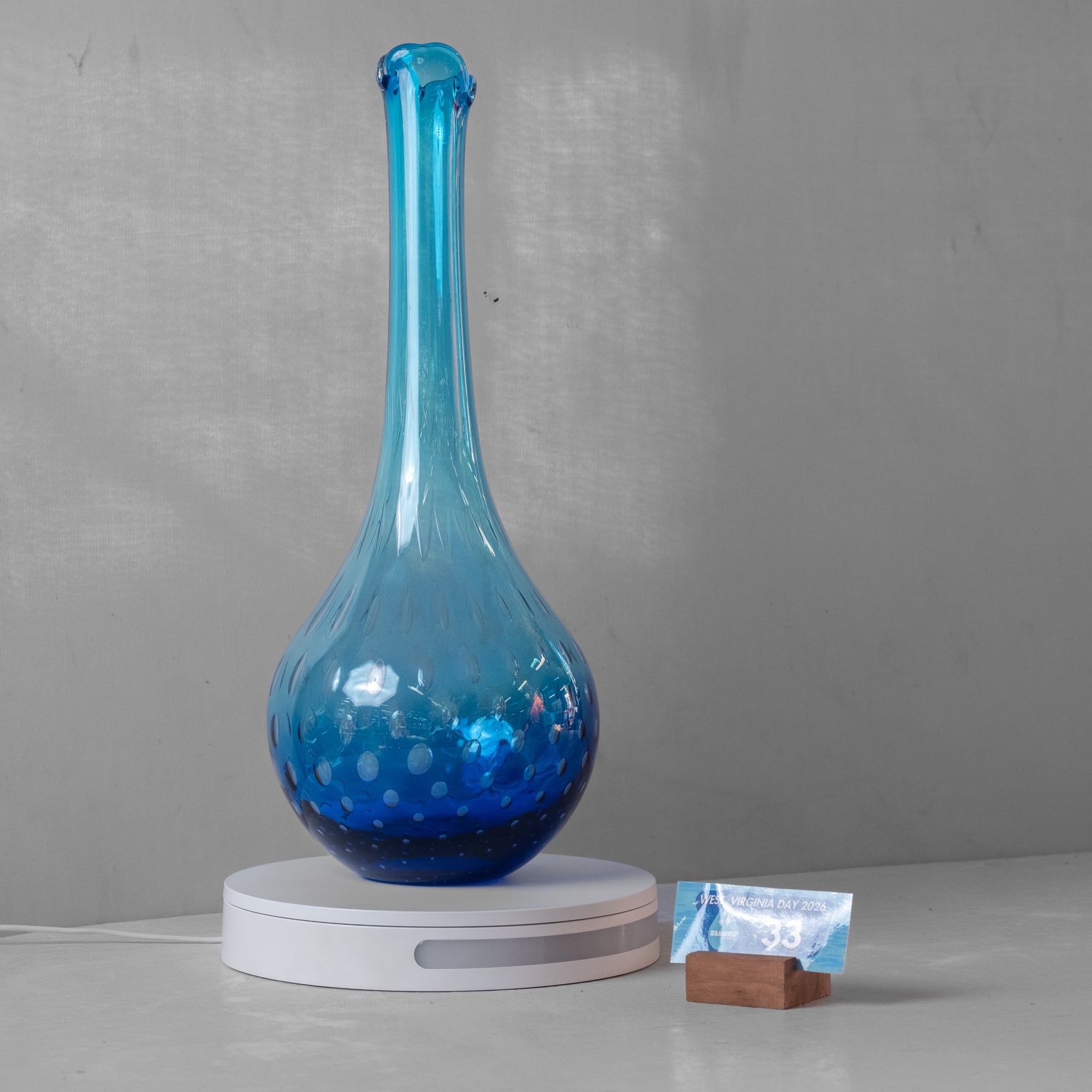 A blue glass vase with a bulbous base and a long, flared neck sits on a white circular charging base against a gray wall. A small packet and wooden block are placed beside the base.