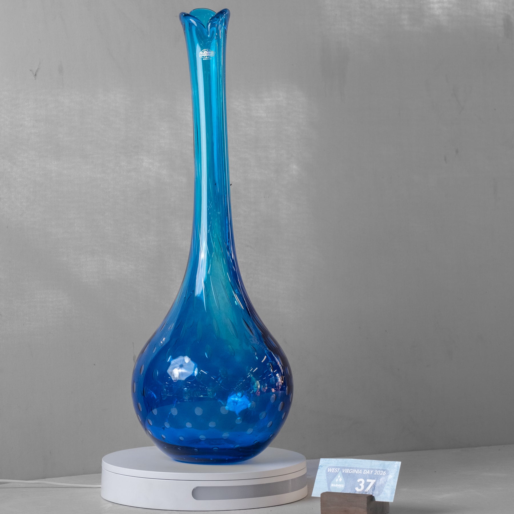 A tall, slender blue glass vase with a flared rim and a bulbous base, displayed on a white circular stand against a light gray wall.