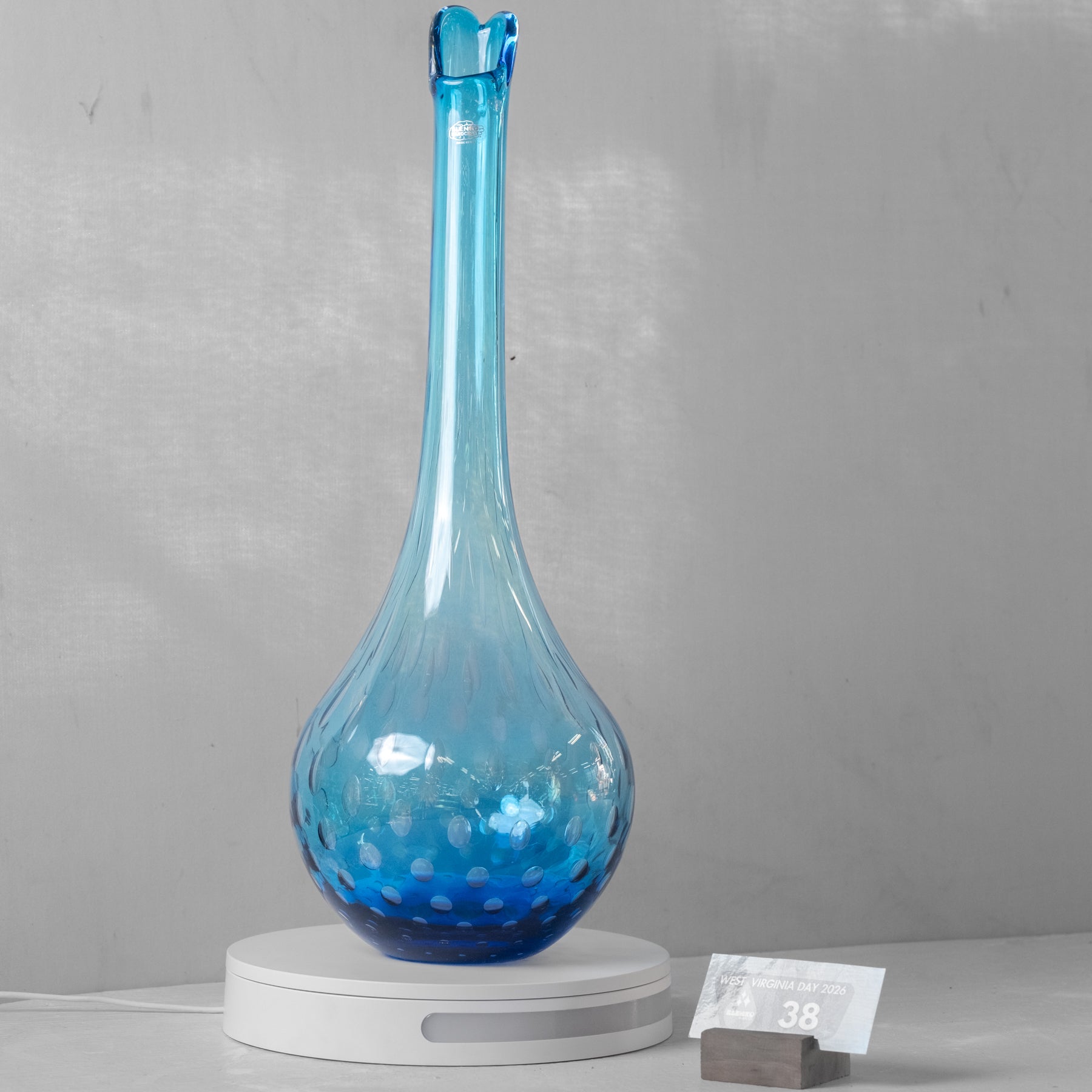 A tall, bulb-shaped blue glass vase with a narrow neck and small flared rim, displayed on a white circular stand against a light gray wall. A small price card is visible beside the stand.