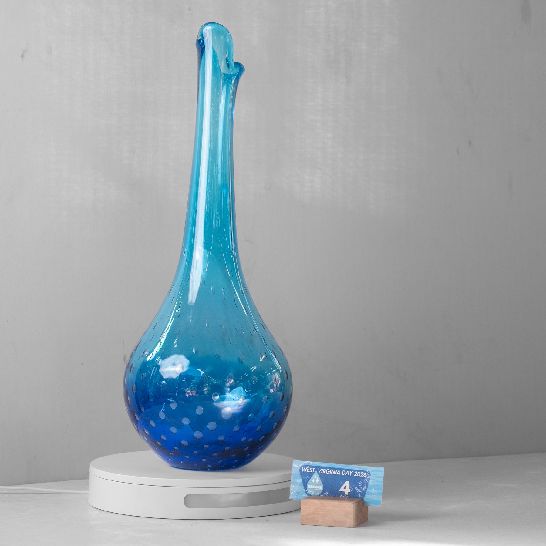 A tall, narrow blue glass vase with a bulbous base and small white speckles, displayed on a white circular pedestal. A small card on a wooden block in front shows a price.